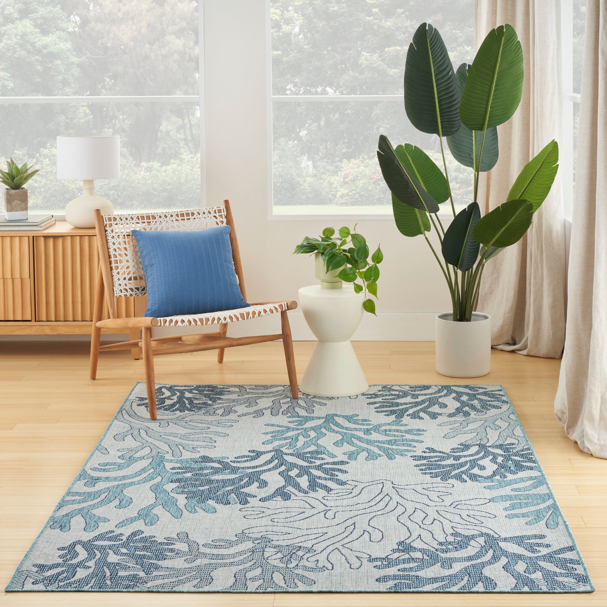 Cozy living room corner with a wooden woven chair, blue cushion, large leafy plant, and blue leaf-pattern rug by a window.