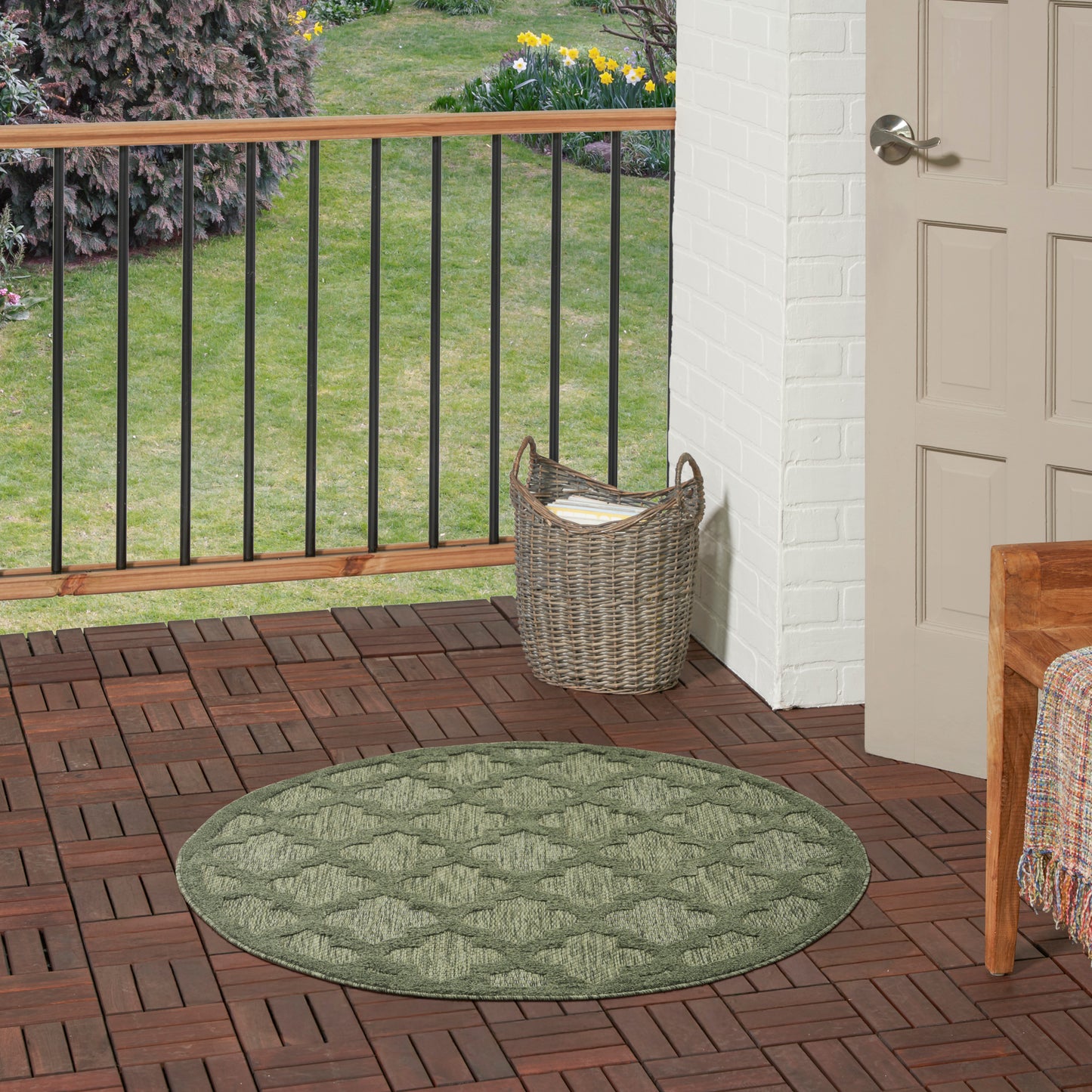 Round green braided outdoor rug on brick porch with wicker basket, white brick wall, wooden railing, and garden beyond.