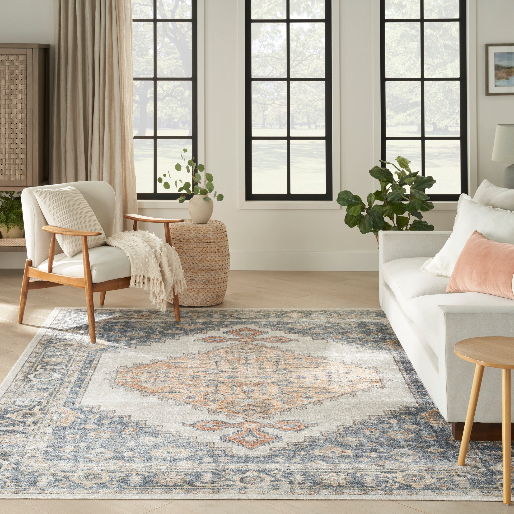 Neutral living room with large blue-gray oriental rug, cream sofa, wooden armchair, round wicker table, and greenery.