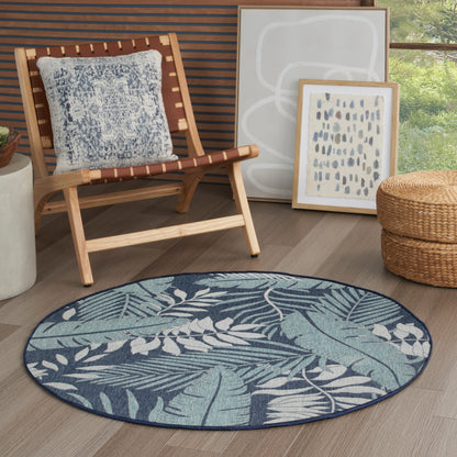 Round teal-blue tropical leaf rug on wood floor beside a wooden sling chair, blue patterned cushion, wicker ottoman, and leaning framed art.