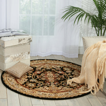 Round rug with black, cream, gold and red floral pattern on light wood floor, beside a beige knit throw and potted plant.