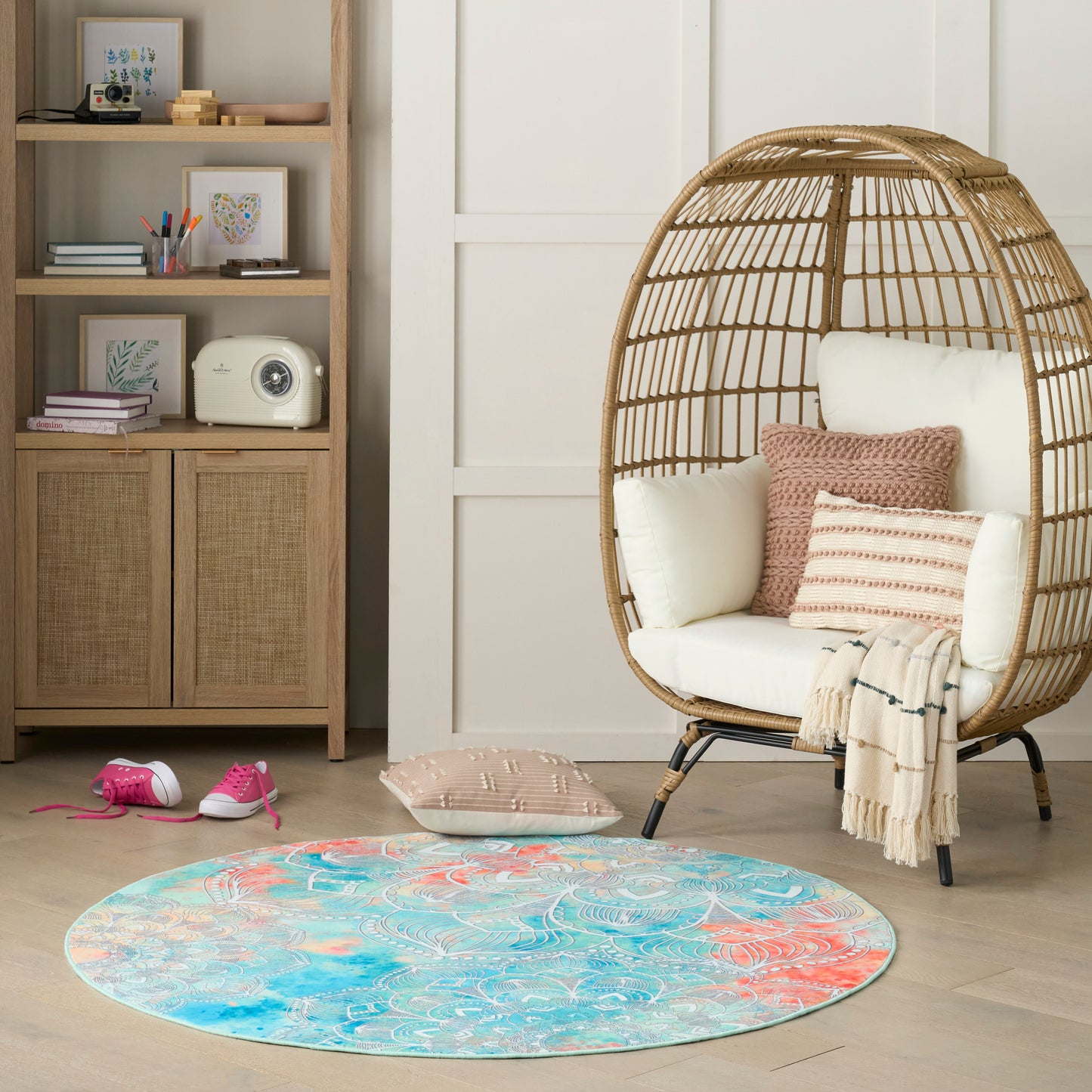Round blue and coral patterned rug beside a white wicker egg chair with cream cushions and pink pillows.