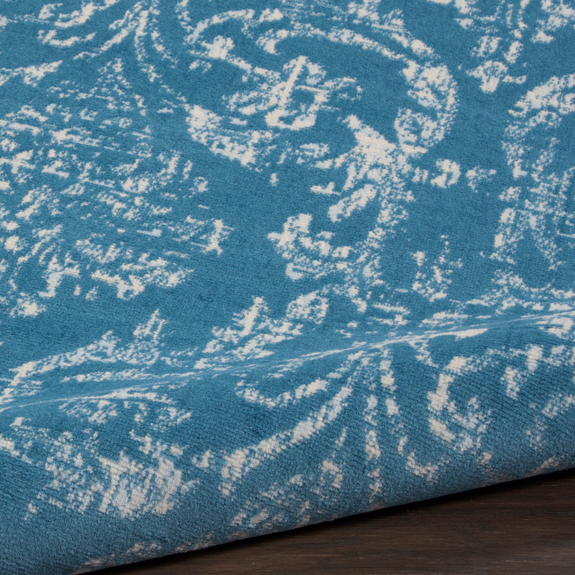 Teal blue damask patterned fabric with white floral motif, folded edge on dark wood surface; great for upholstery or drapery.