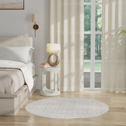 Neutral bedroom with white bedding, round white nightstand, curved glass lamp, books, beige curtains, window, circular rug.