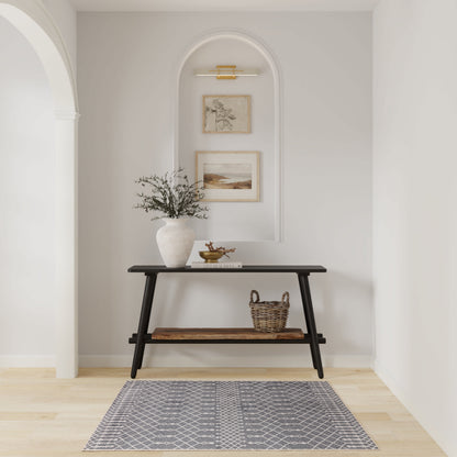 Black wooden two-tier console table in a pale entry alcove with a vase of greenery, woven basket, arch niche with framed art, and a grey patterned rug.