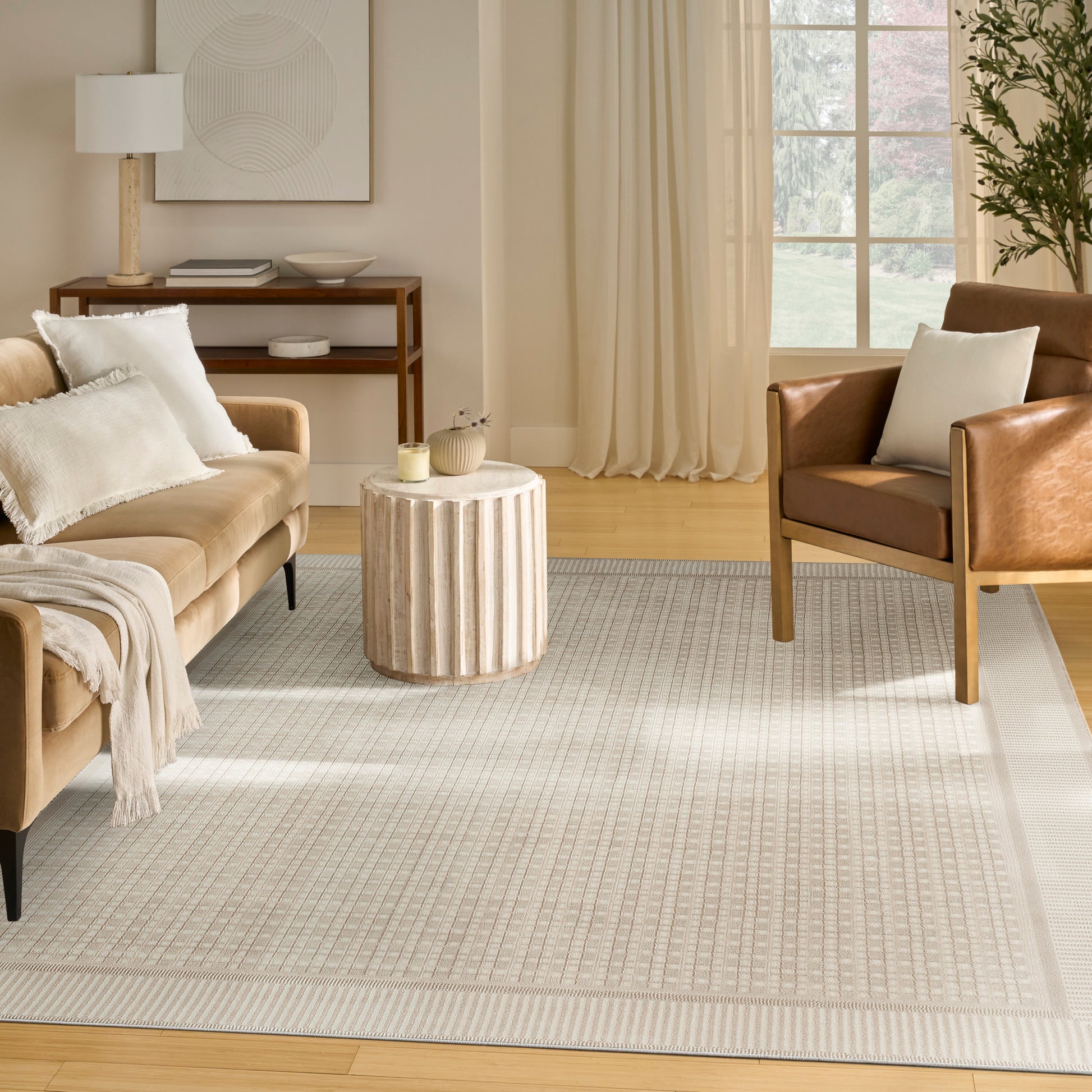 Neutral living room featuring a tan sofa, a brown leather chair, a ribbed round wooden side table, a light gray patterned rug, and sheer curtains.