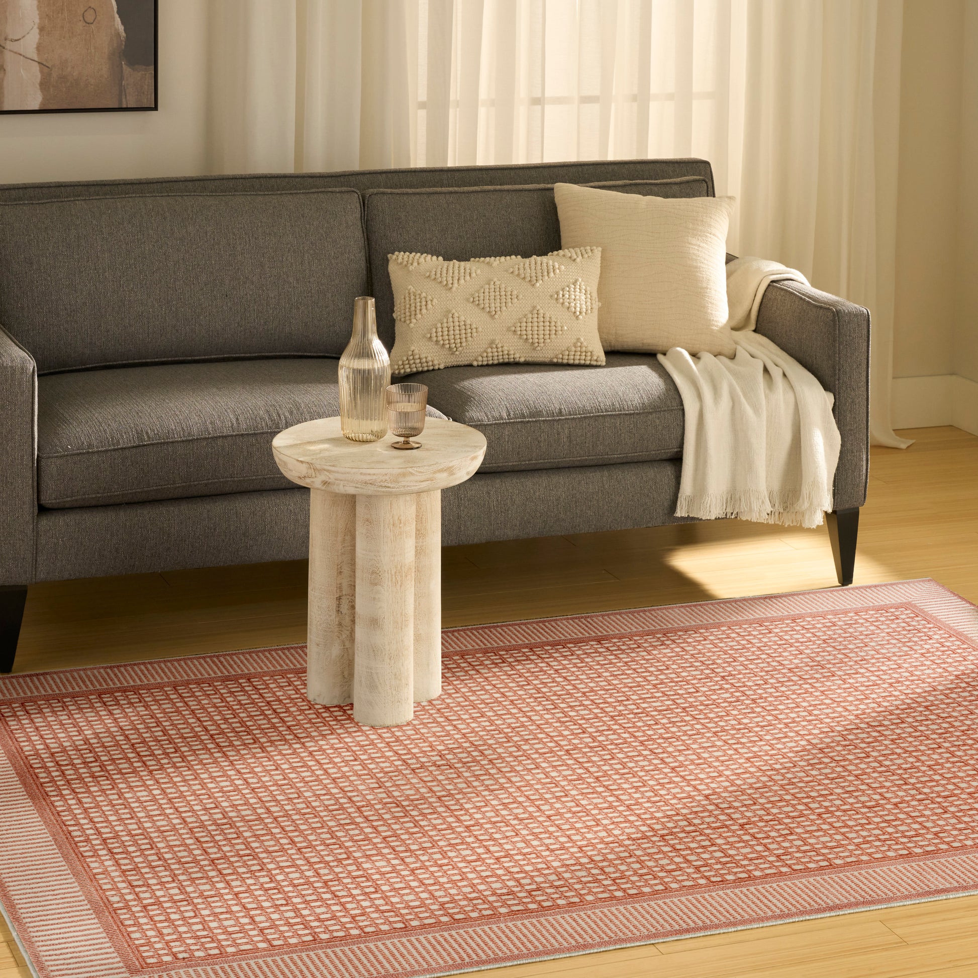 Grey fabric sofa with textured cream cushions and throw, round light wood side table, pink geometric rug in a living room.
