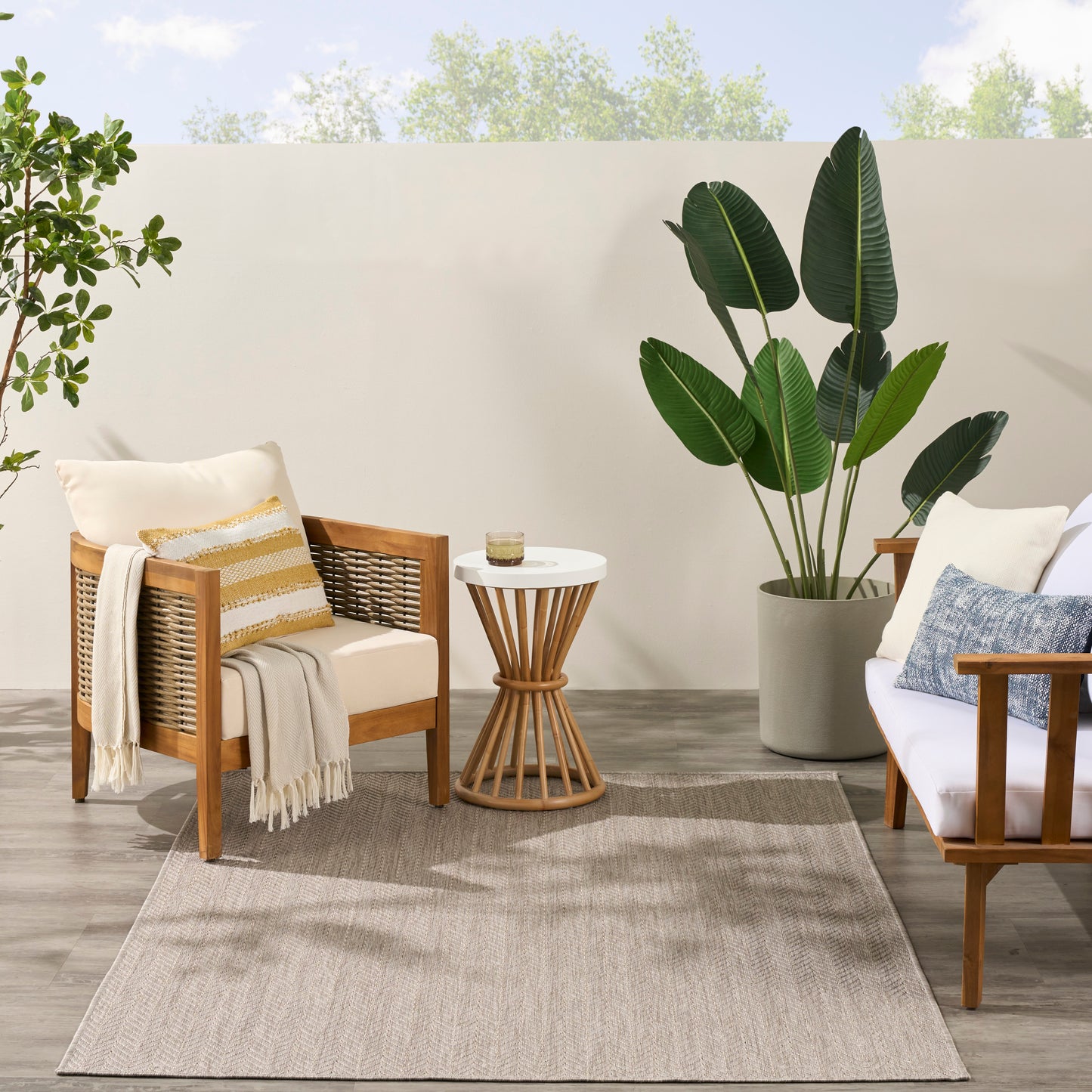 Outdoor seating with wooden frames, woven backs, beige cushions, striped pillow, throw, round white-top side table, plant.