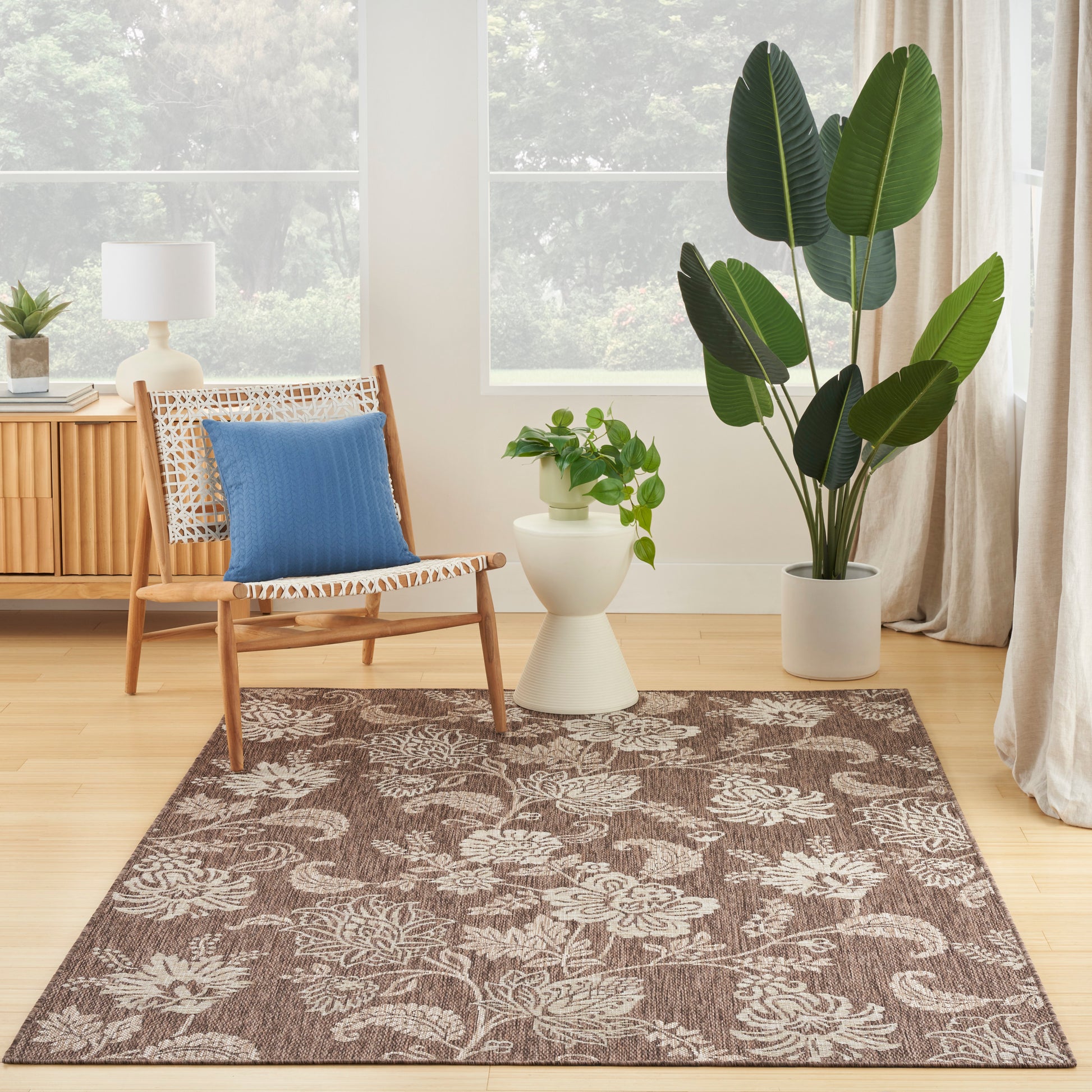Brown floral area rug with beige leaves, wooden chair with blue cushion, potted plants, cream side table in living room.