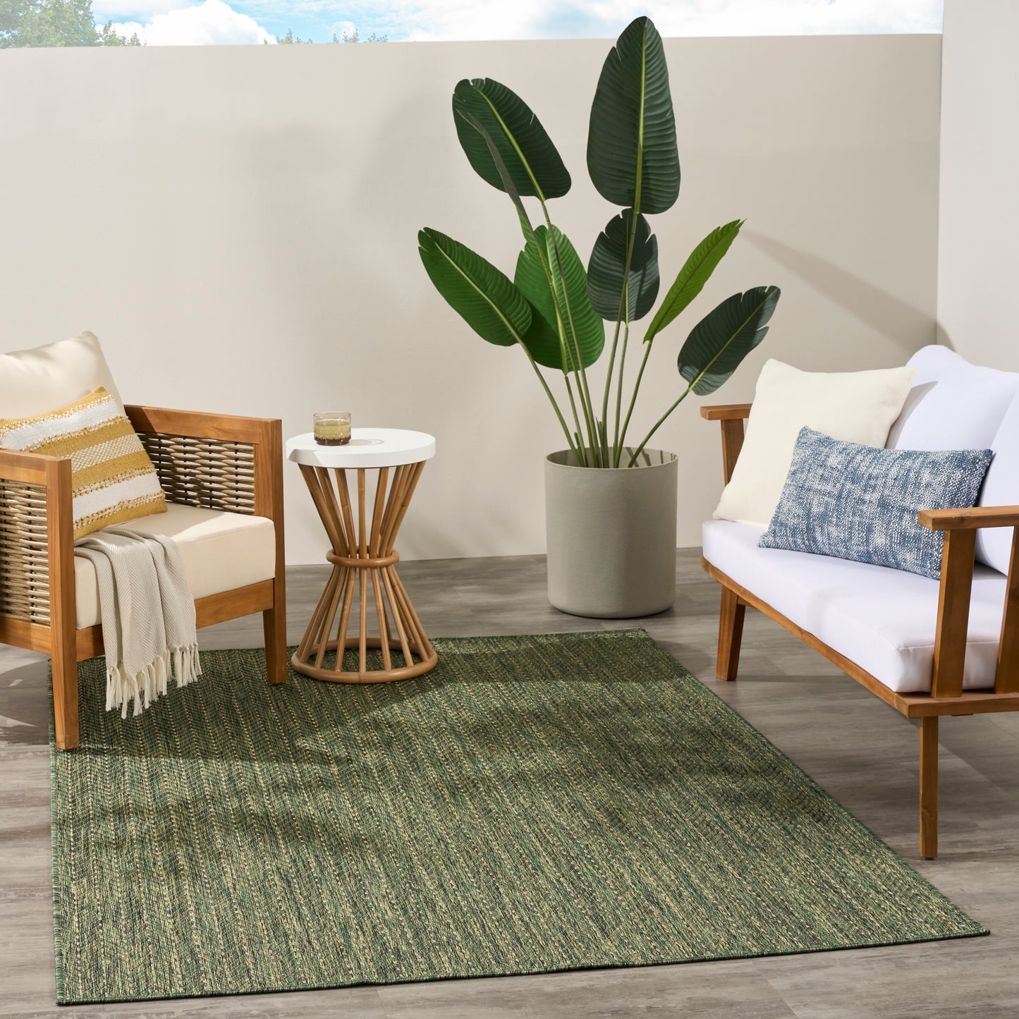Outdoor patio seating with wooden frames, white cushions, beige throw, white table, large potted tropical plant, green rug.