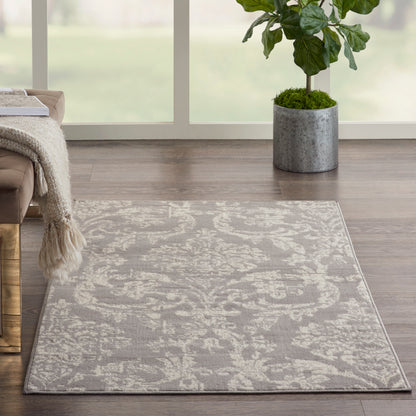 Grey patterned area rug with subtle floral motif on dark wood floor; potted plant in concrete pot by sunny window.