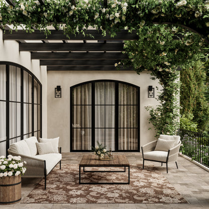 Cozy outdoor patio with cream cushions, black arched doors, wooden coffee table, and a brown floral rug under a pergola.