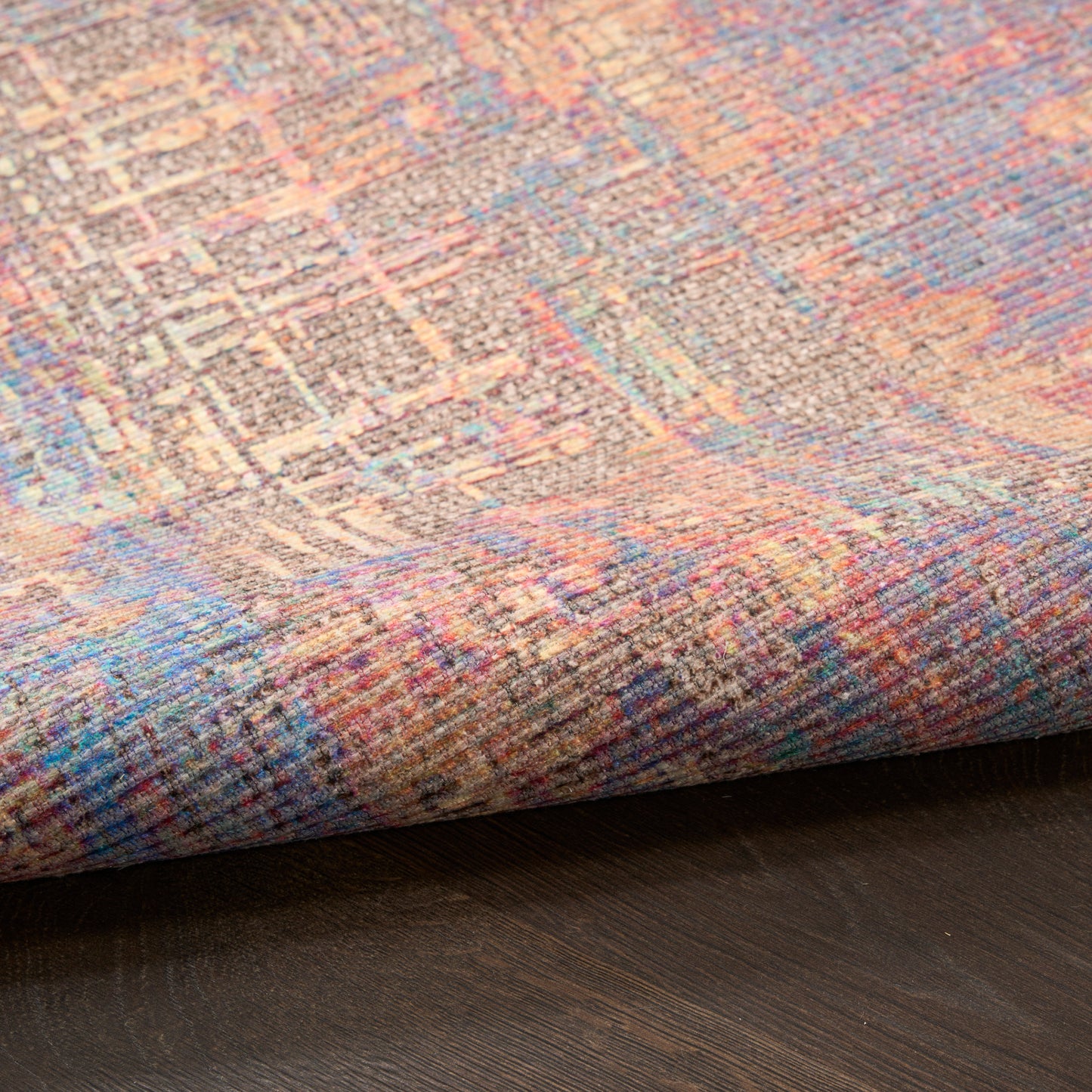 Close-up of a multicolored woven rug with pastel pink, blue and lavender tones, folded edge on dark wood floor.