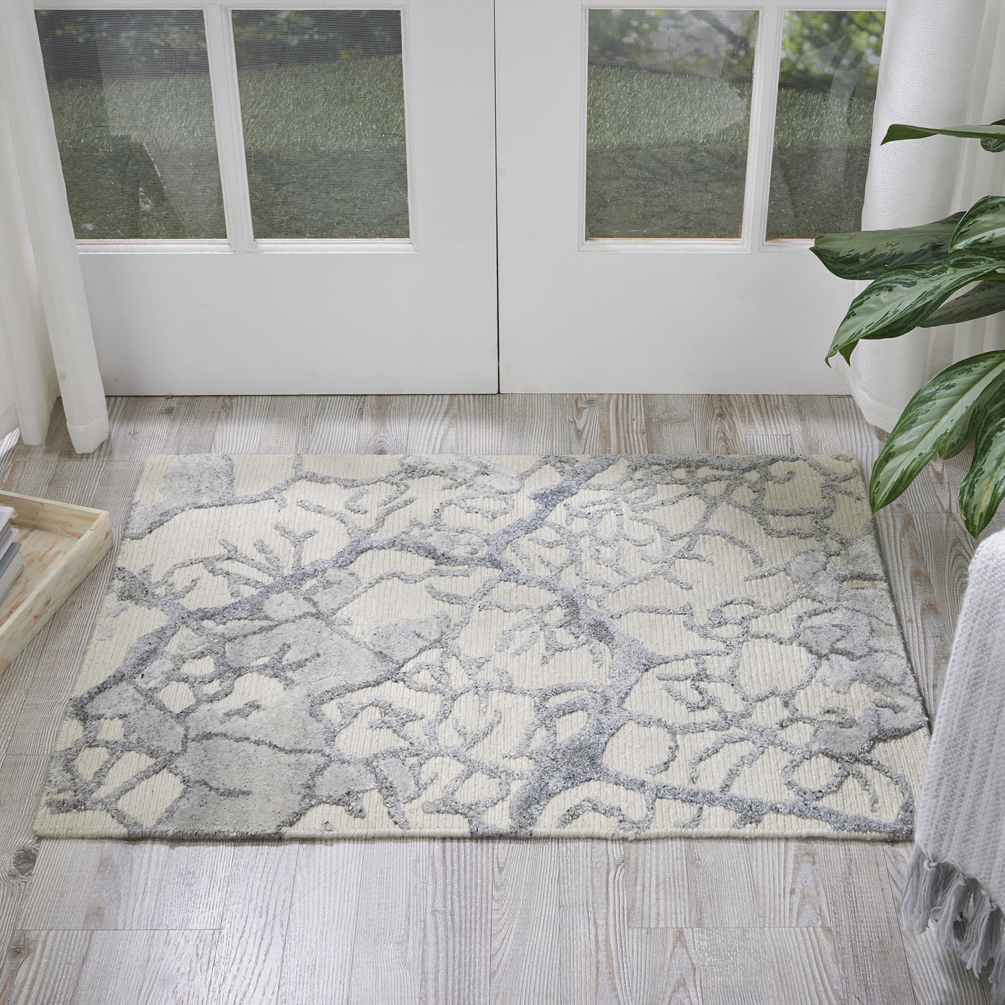 Rectangular cream area rug with grey abstract branch pattern on light wood floor by glass-paneled double doors.
