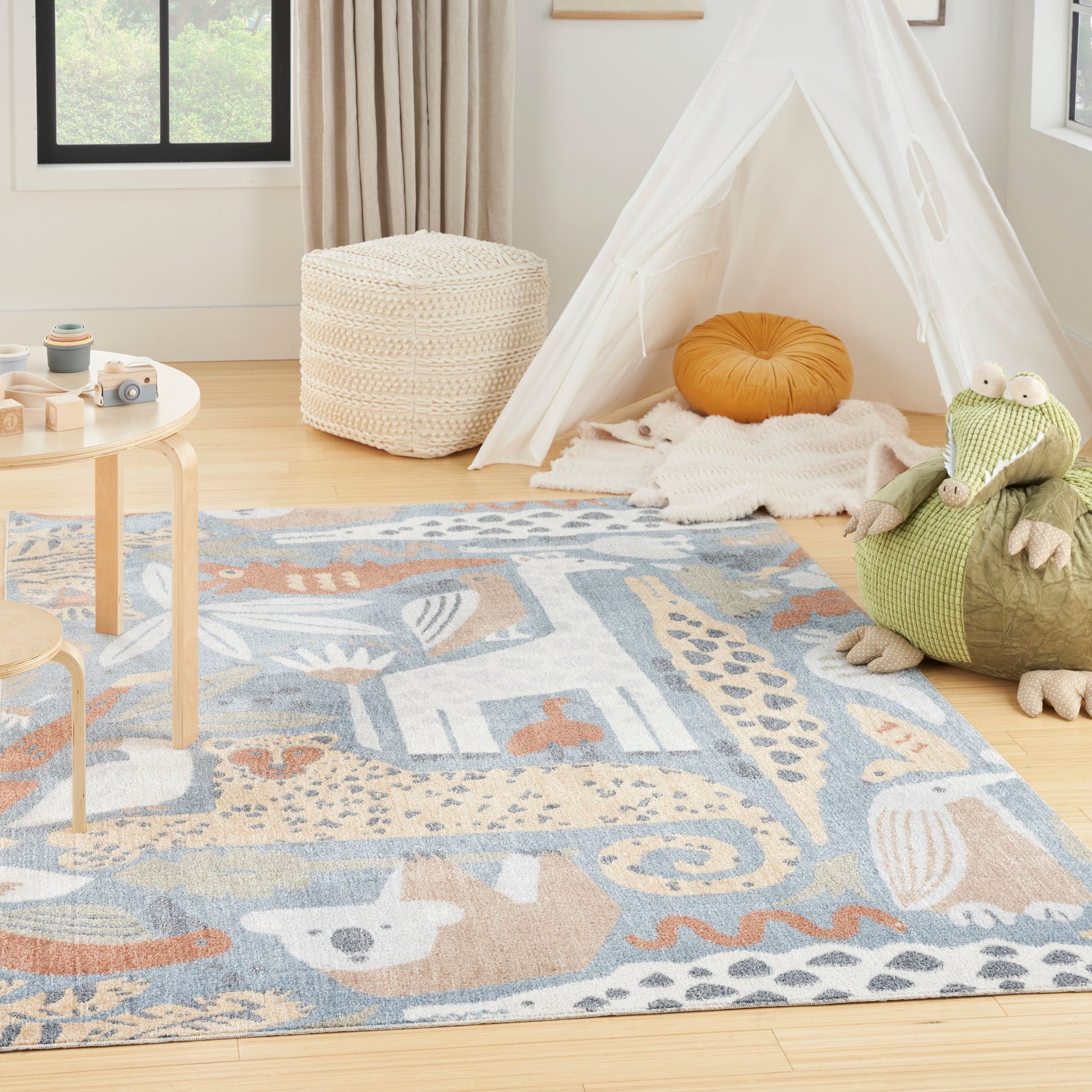 Kids' rug with an animal design in a playroom, alongside a white teepee tent with sheer panels, an orange cushion, a woven pouf, and a green plush crocodile.