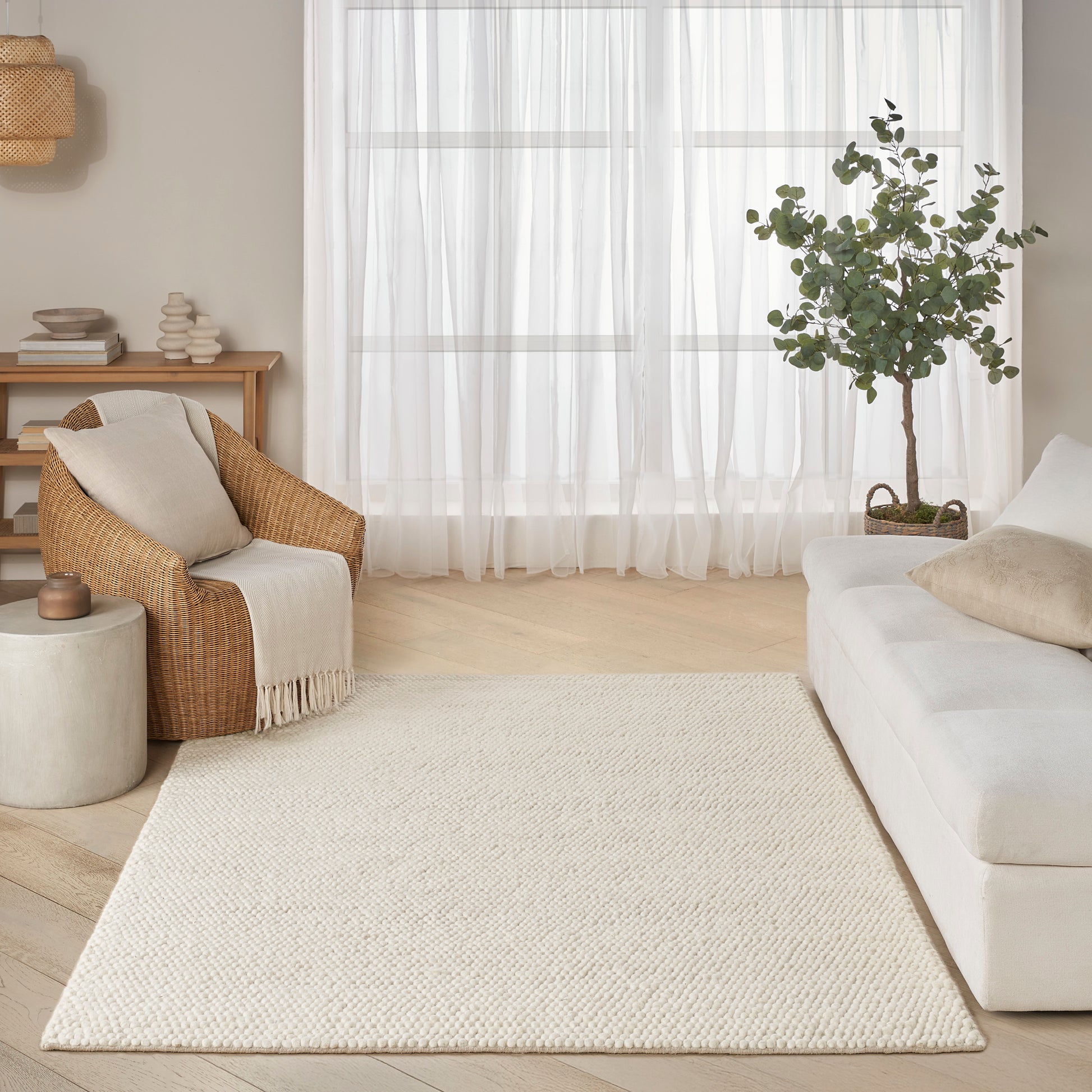 Cream woven area rug on light wood floor beside a white sofa, wicker chair, sheer curtains and a potted tree.