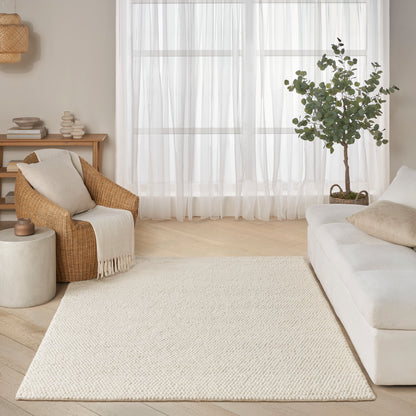 Cream woven area rug on light wood floor beside a white sofa, wicker chair, sheer curtains and a potted tree.