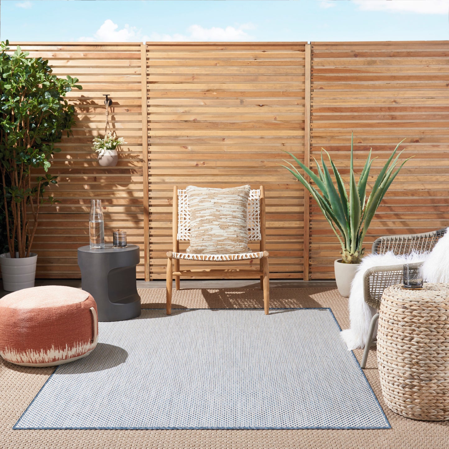 Outdoor boho patio with slat fence, rattan chair, cream cushion, orange pouf, blue-gray rug, wicker table, potted plants.