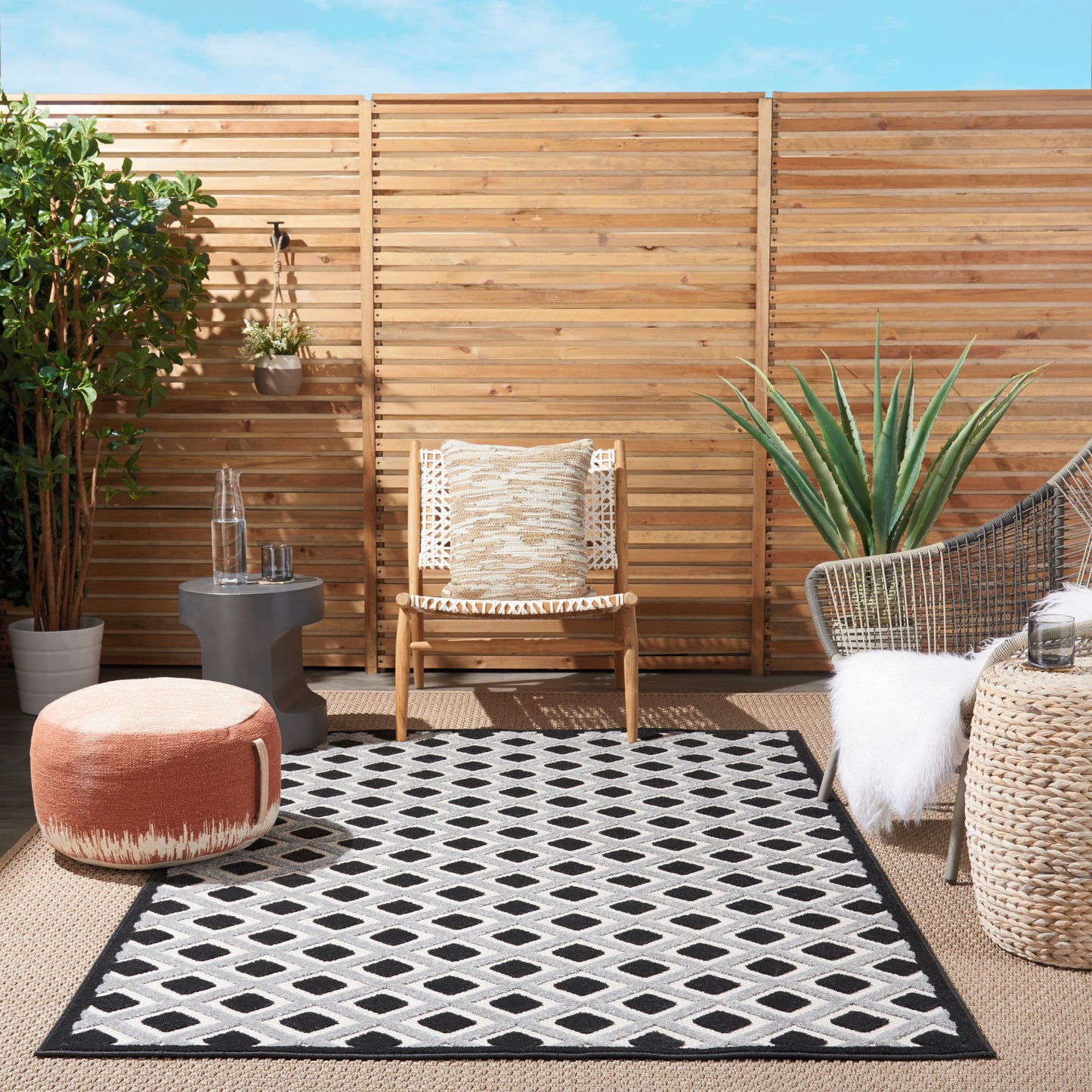 Outdoor patio with wooden slat fence, wicker chairs, black-and-white geometric rug, orange pouf, agave plant.