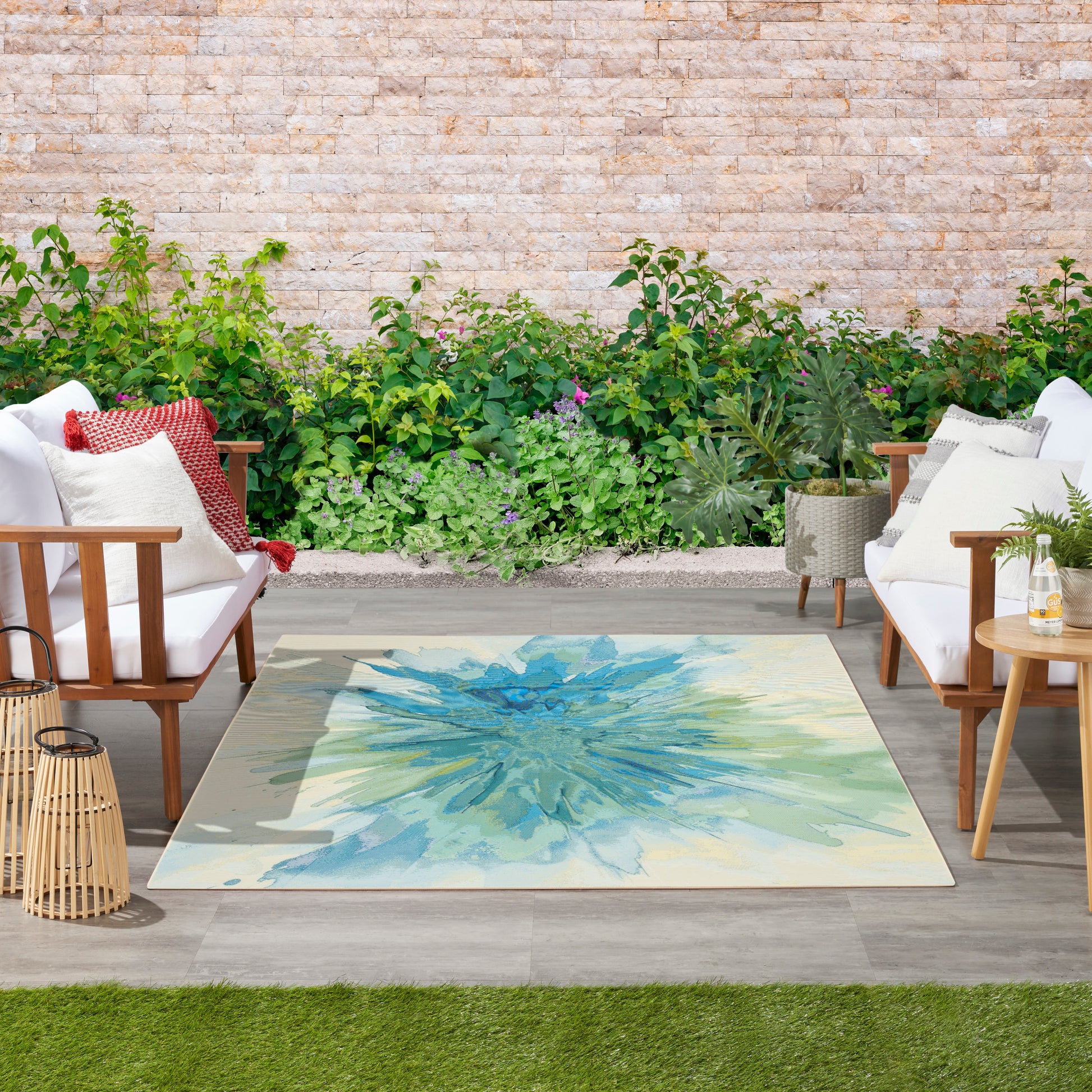 Outdoor lounge with white cushioned wooden chairs, a blue-green abstract rug, lanterns, and greenery against a stone wall.