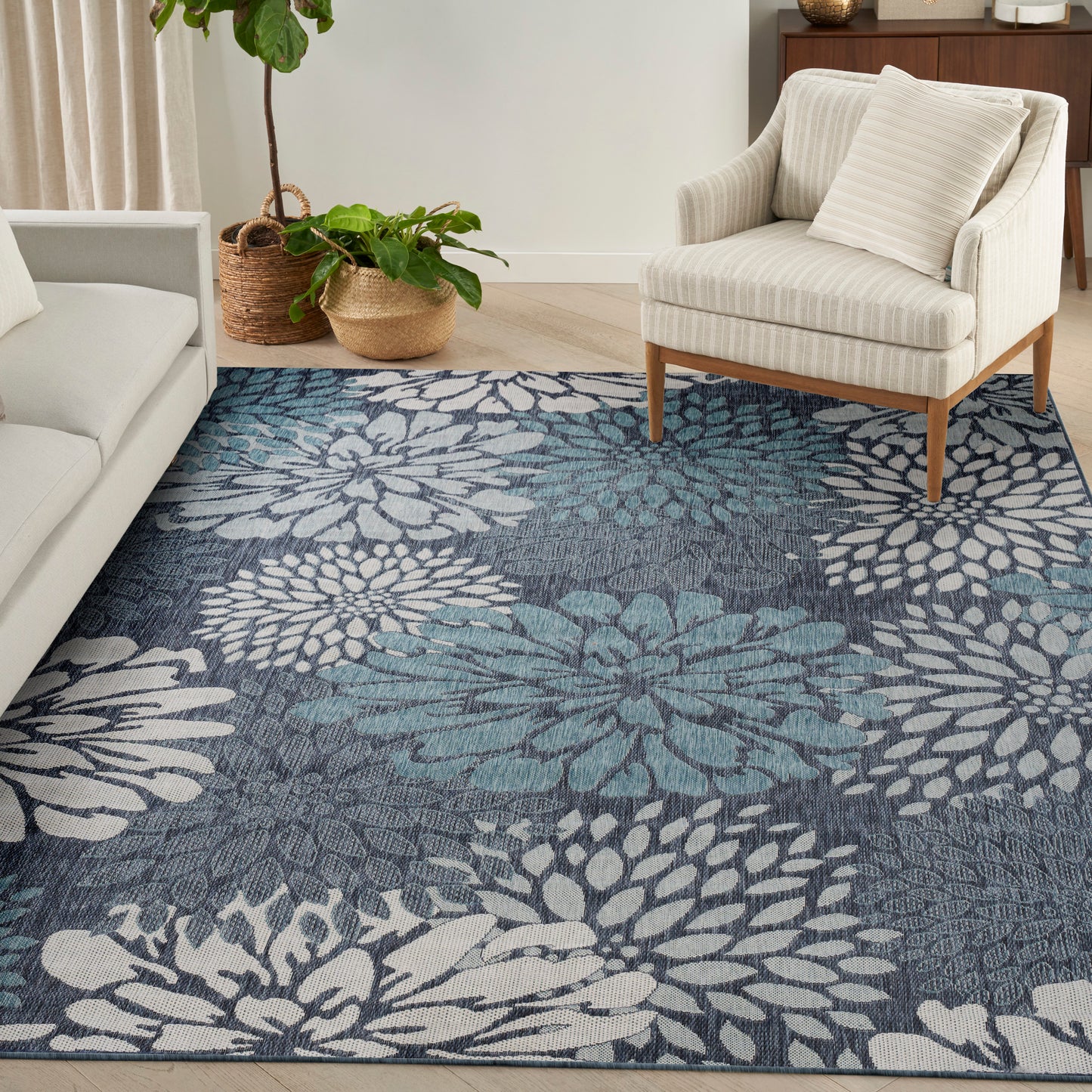 Cream sofa & striped beige armchair on a blue tropical rug with large white petals; potted plants in wicker baskets.