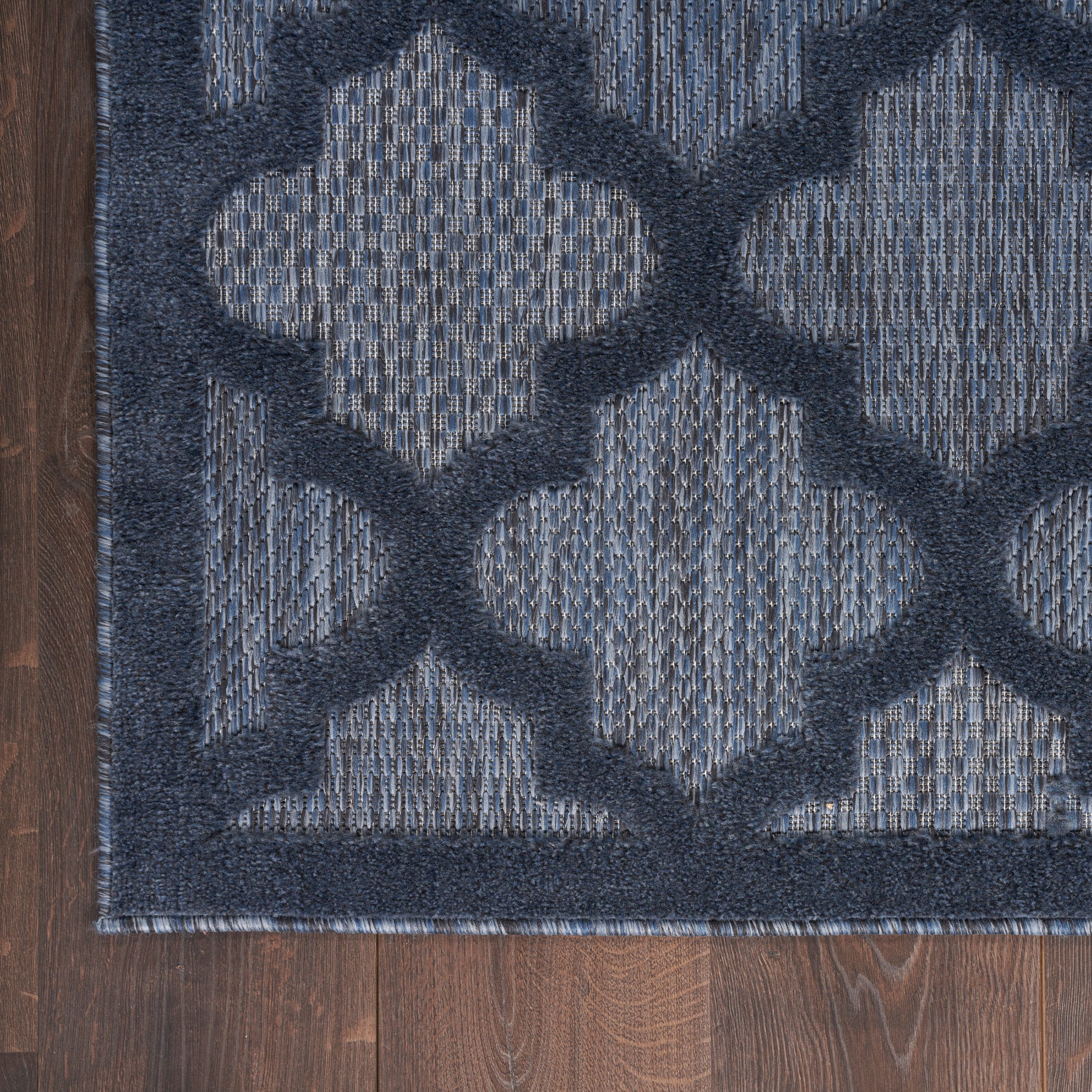 Blue geometric trellis rug with light blue quatrefoil motifs on a dark navy background, woven texture on wood floor.