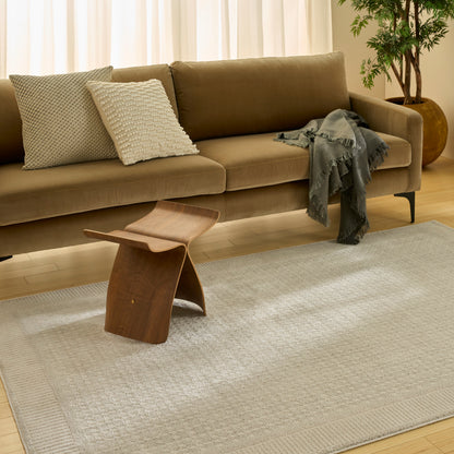 Taupe fabric sofa with textured cushions, gray fringe throw, curved wooden side stool, beige rug, potted plant corner