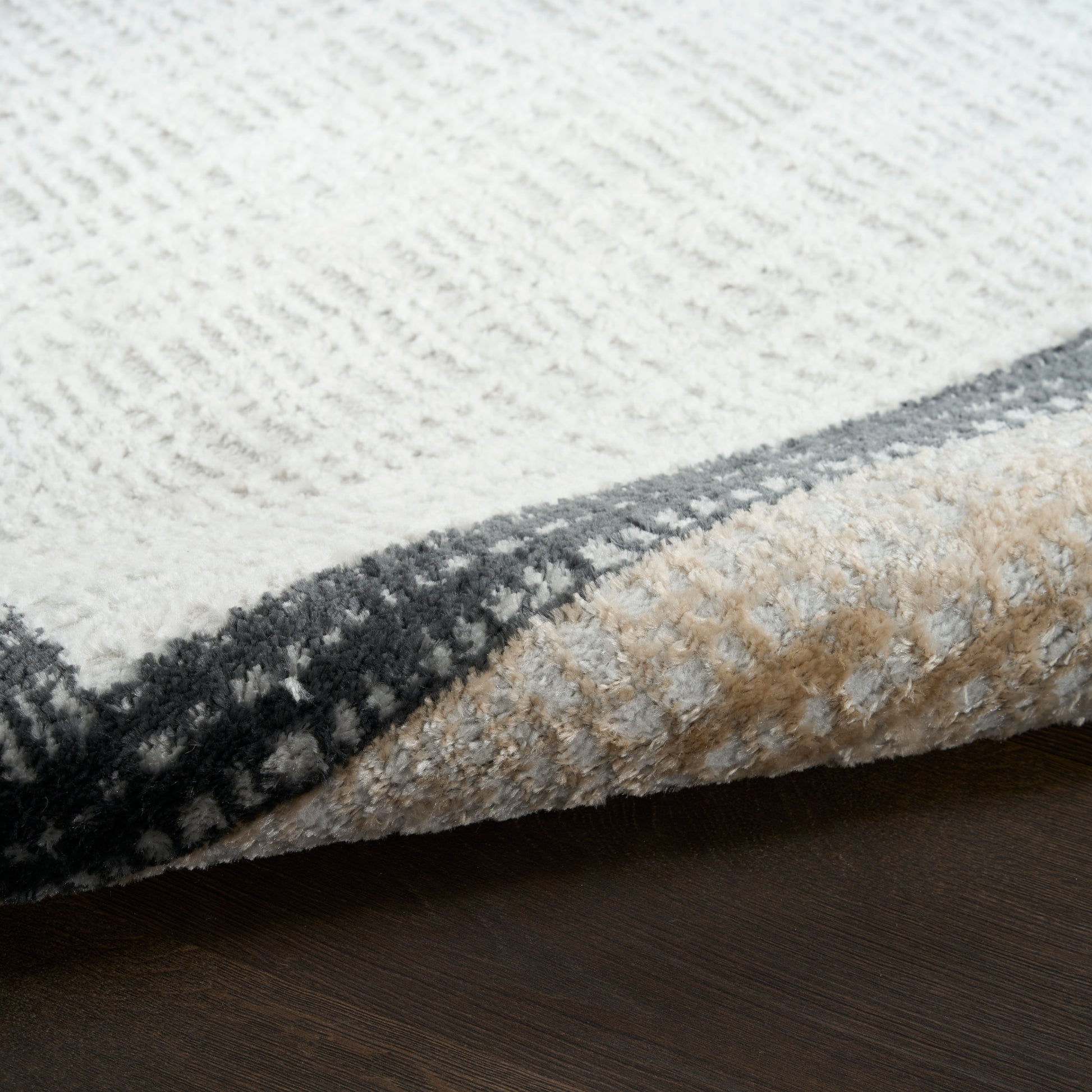 Close-up of a textured cream and gray geometric area rug with a folded beige underside on dark wood floor.
