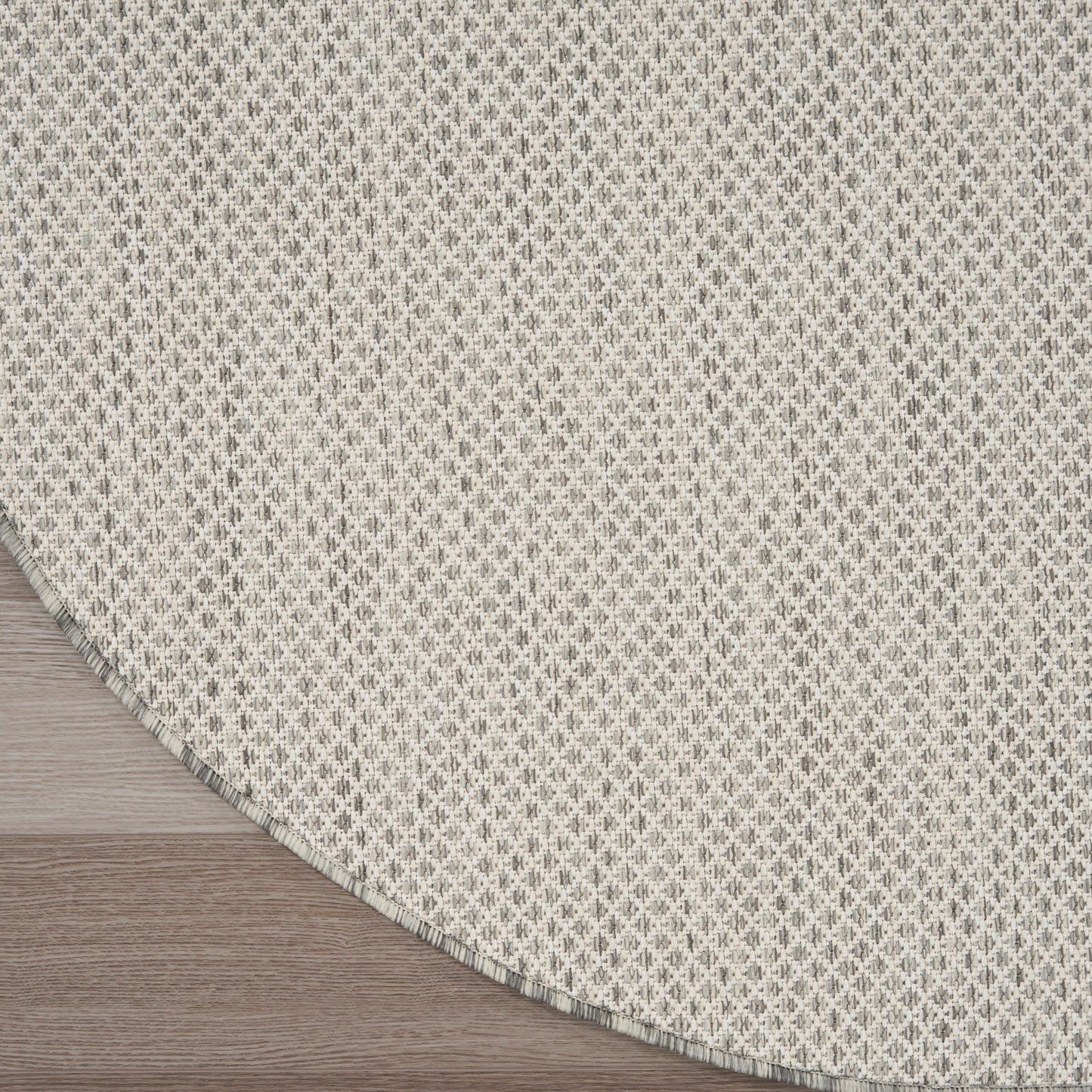 Round beige woven rug with small repeating geometric pattern and curved edge on light wood floor.