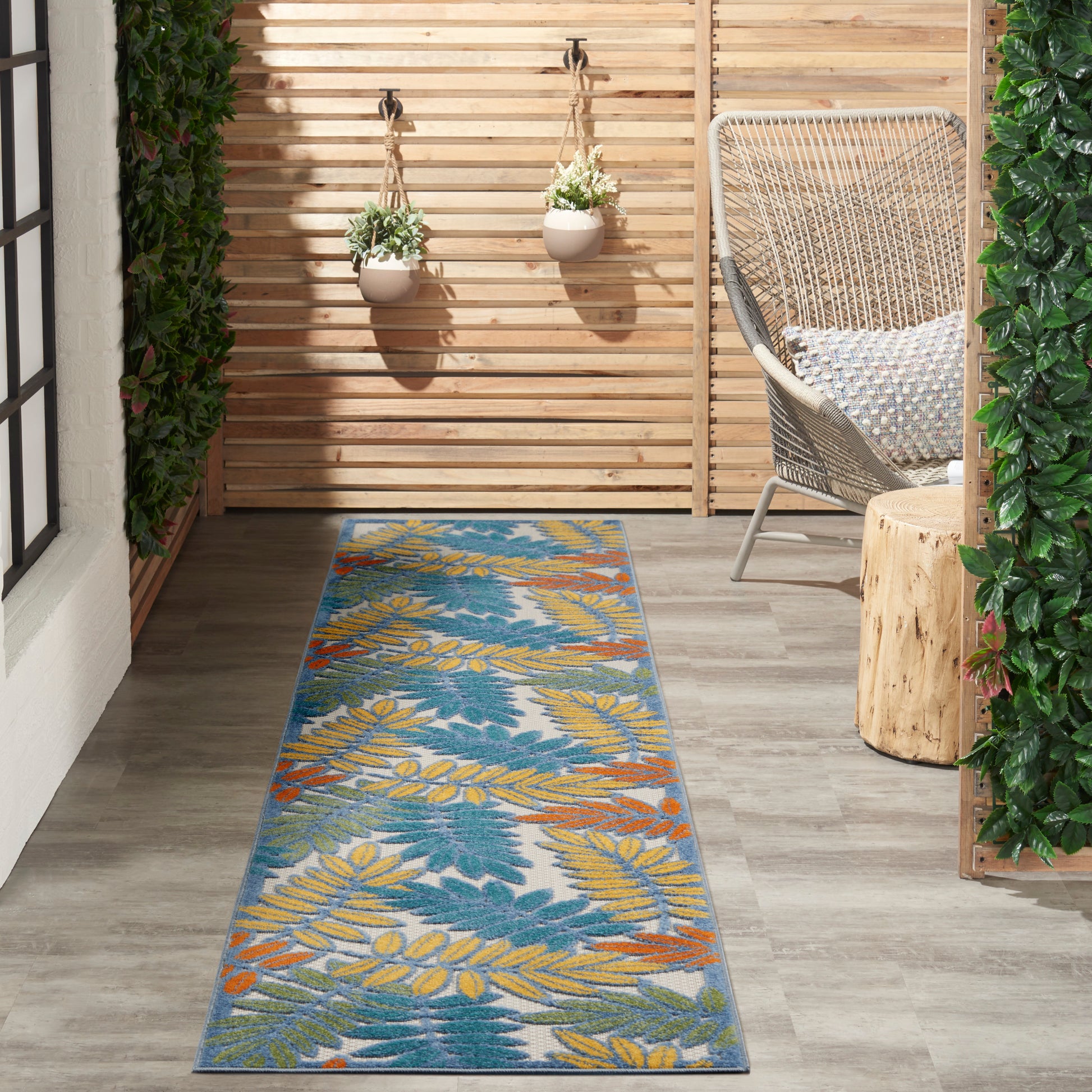 Outdoor rug runner with blue background and yellow-leaf print beside a wicker chair, wooden slat fence and hanging planters.