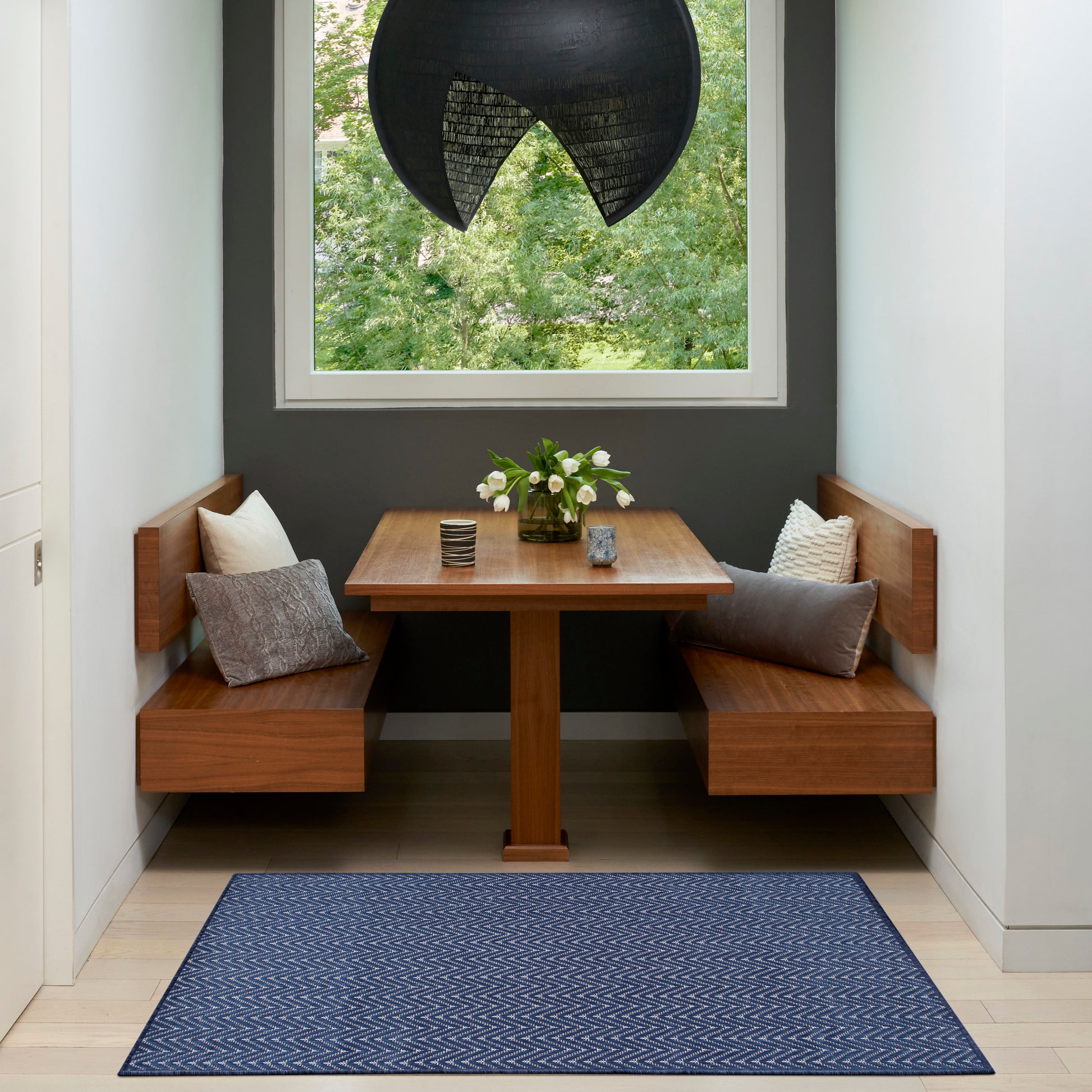 Built-in wooden dining nook with two benches, square table, navy chevron rug, black pendant lamp, and flowers by window.