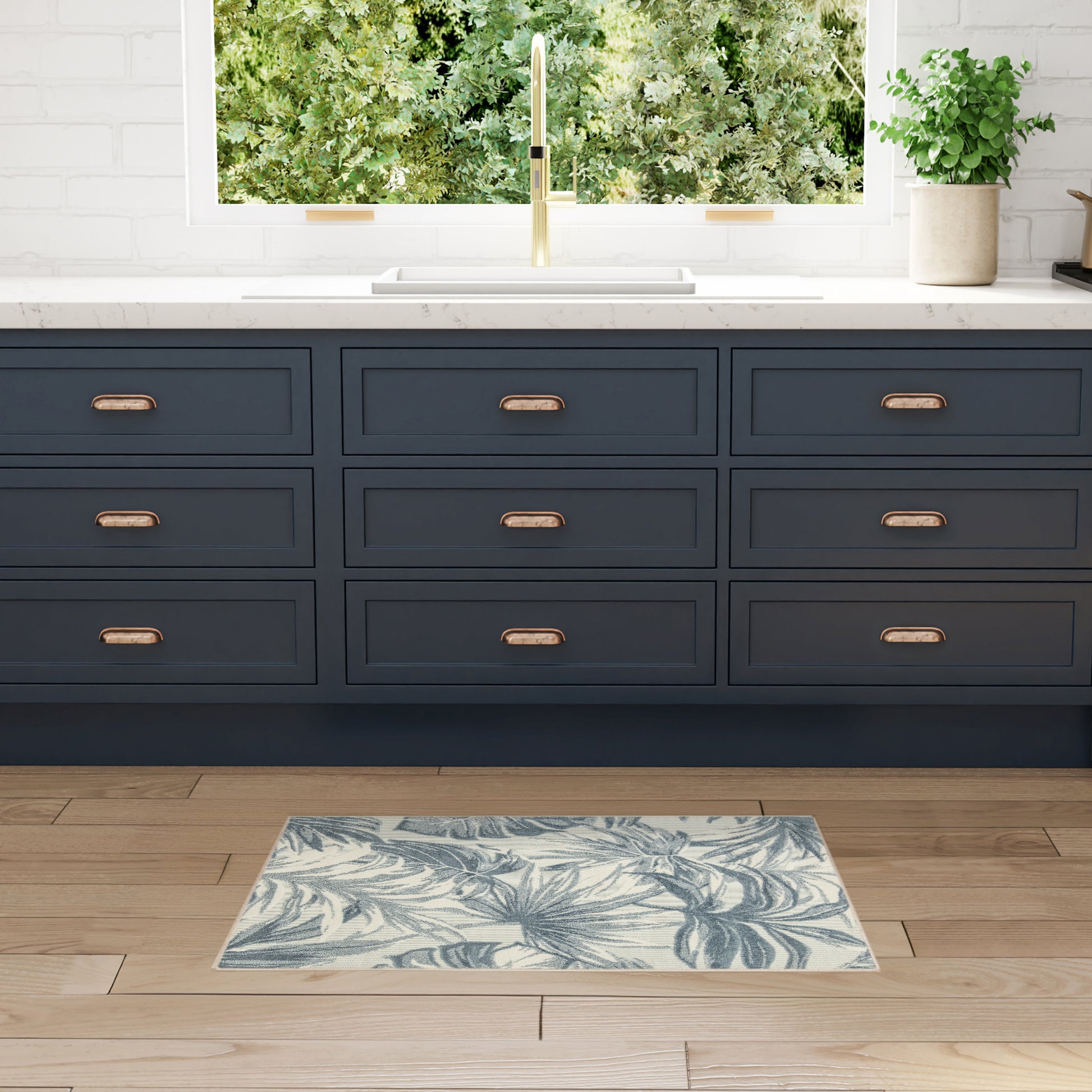 Dark navy cabinets with copper pulls, white countertop, brass faucet, potted plant, and blue palm-leaf rug on wood floor.