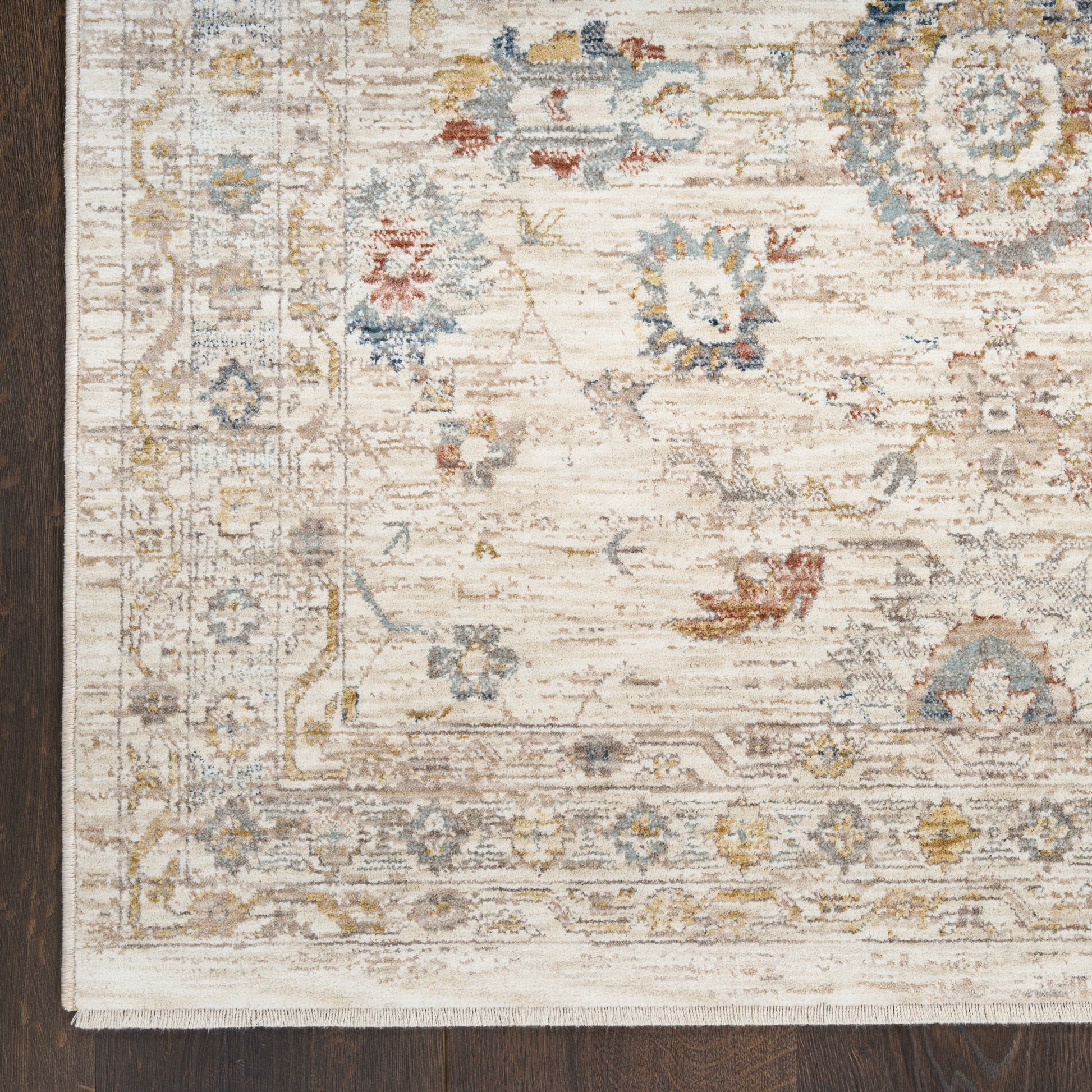 Beige oriental rug with faded blue, yellow and gray florals and scroll border on dark wood floor; fringe at edge.