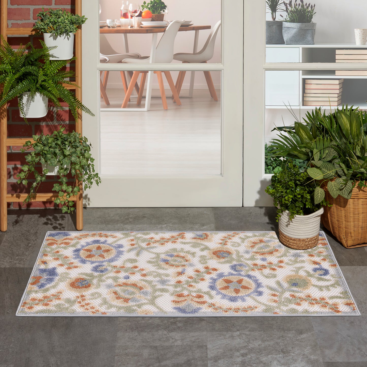 Colorful floral medallion rug outside glass doors; potted plants flank doorway; dining room with white chairs visible inside.