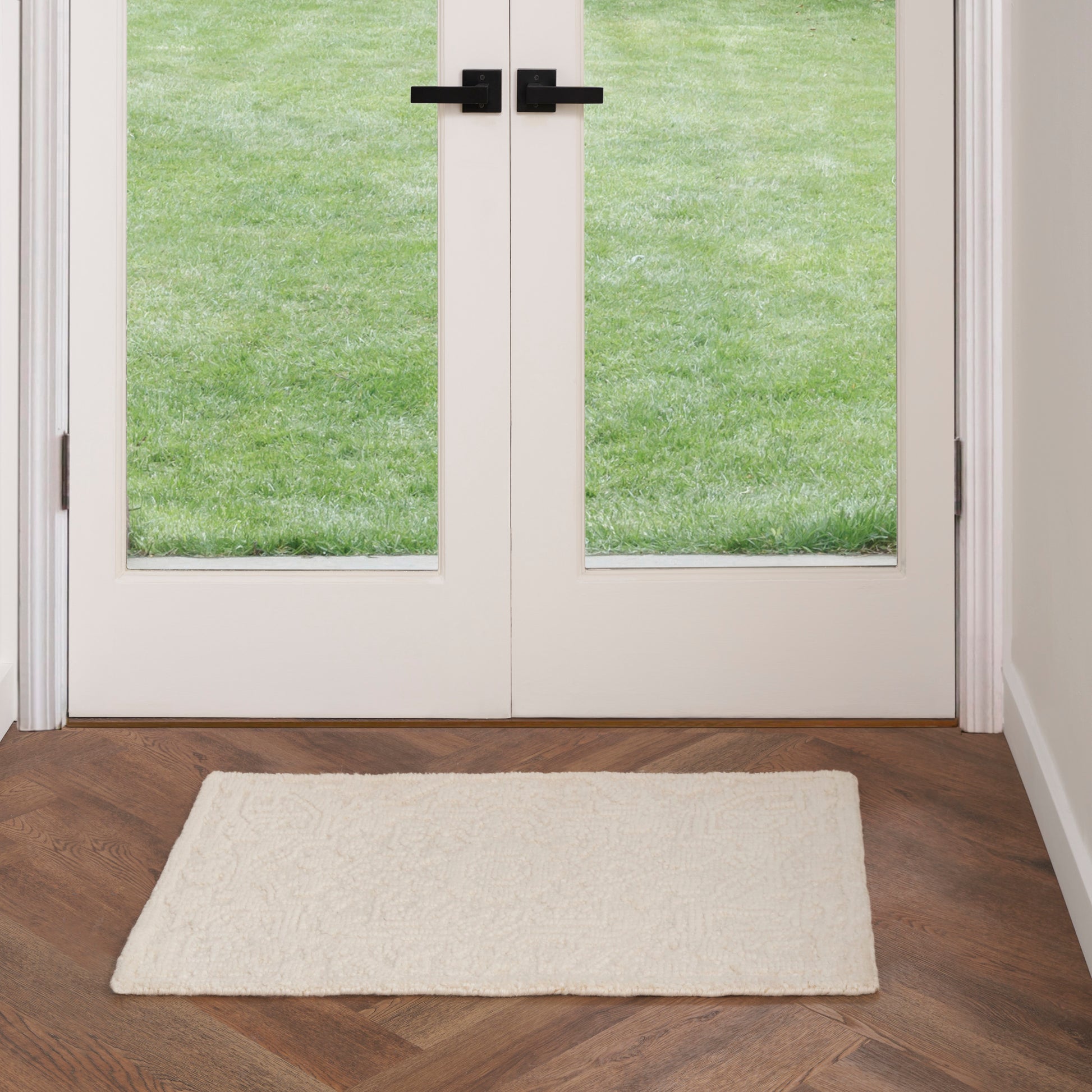 Cream plush entry mat on dark wood herringbone floor in front of white double doors with glass panes and black handles.