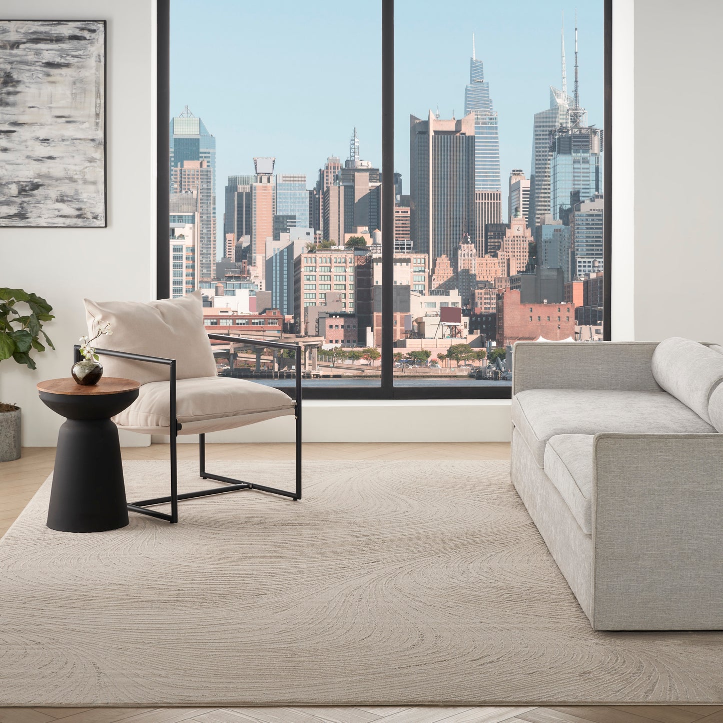 Neutral living room with a beige armchair on a black metal frame, a light rug, a gray sofa, and city skyline visible through large windows.
