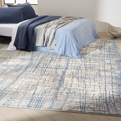 Queen bed with layered navy & light-blue bedding & beige throw & blue abstract-pattern rug.