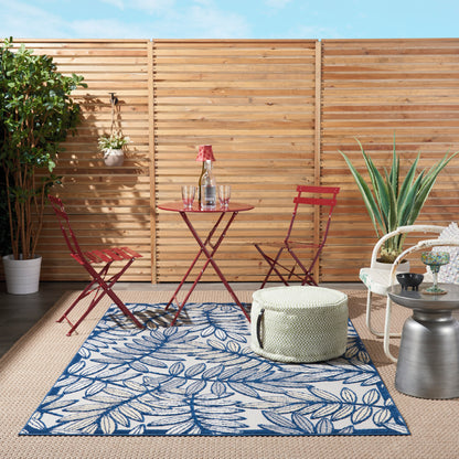 Outdoor patio with blue leaf-pattern rug, red metal folding chairs, small table, potted plants, white wicker chair, and pouf.