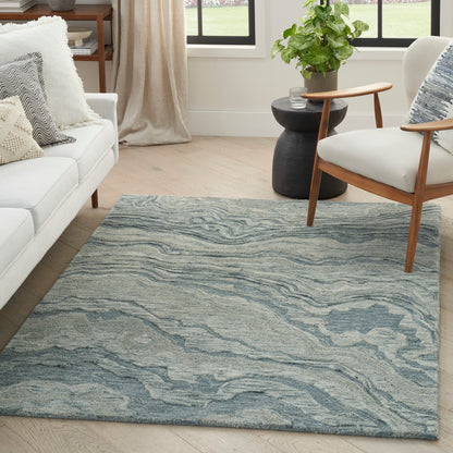 Blue-gray abstract area rug on light hardwood floor in a modern living room with white sofa, wooden armchair, and a plant.