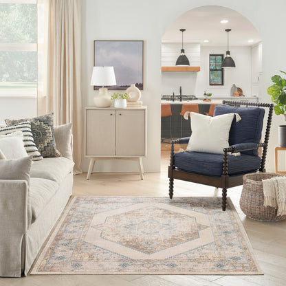Bright living room with a beige fabric sofa, cream cabinet, navy wood-frame armchair, pastel medallion rug, woven basket, and open kitchen arch.