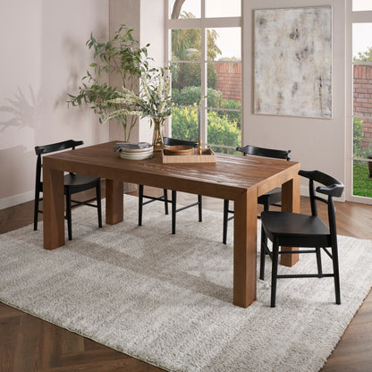 Warm brown rectangular solid wood dining table & thick legs, black chairs, greenery centerpiece on a beige shag rug.
