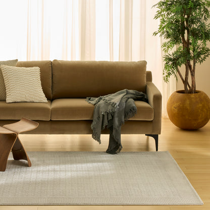 Beige fabric sofa with white textured pillow, gray fringed throw, curved wooden side table, light gray rug, potted tree.