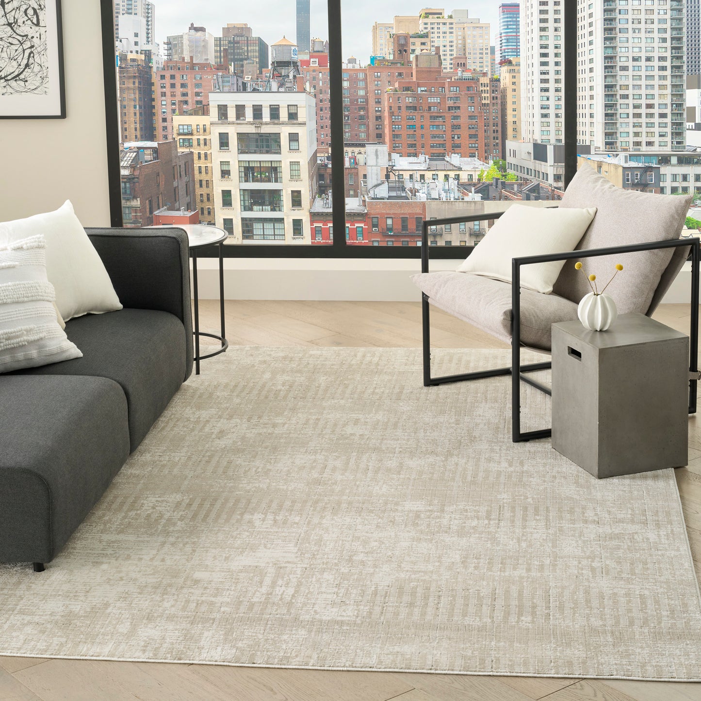 Modern living room with beige rug, dark gray sofa, metal-frame chairs, gray side table with vase, city skyline view.