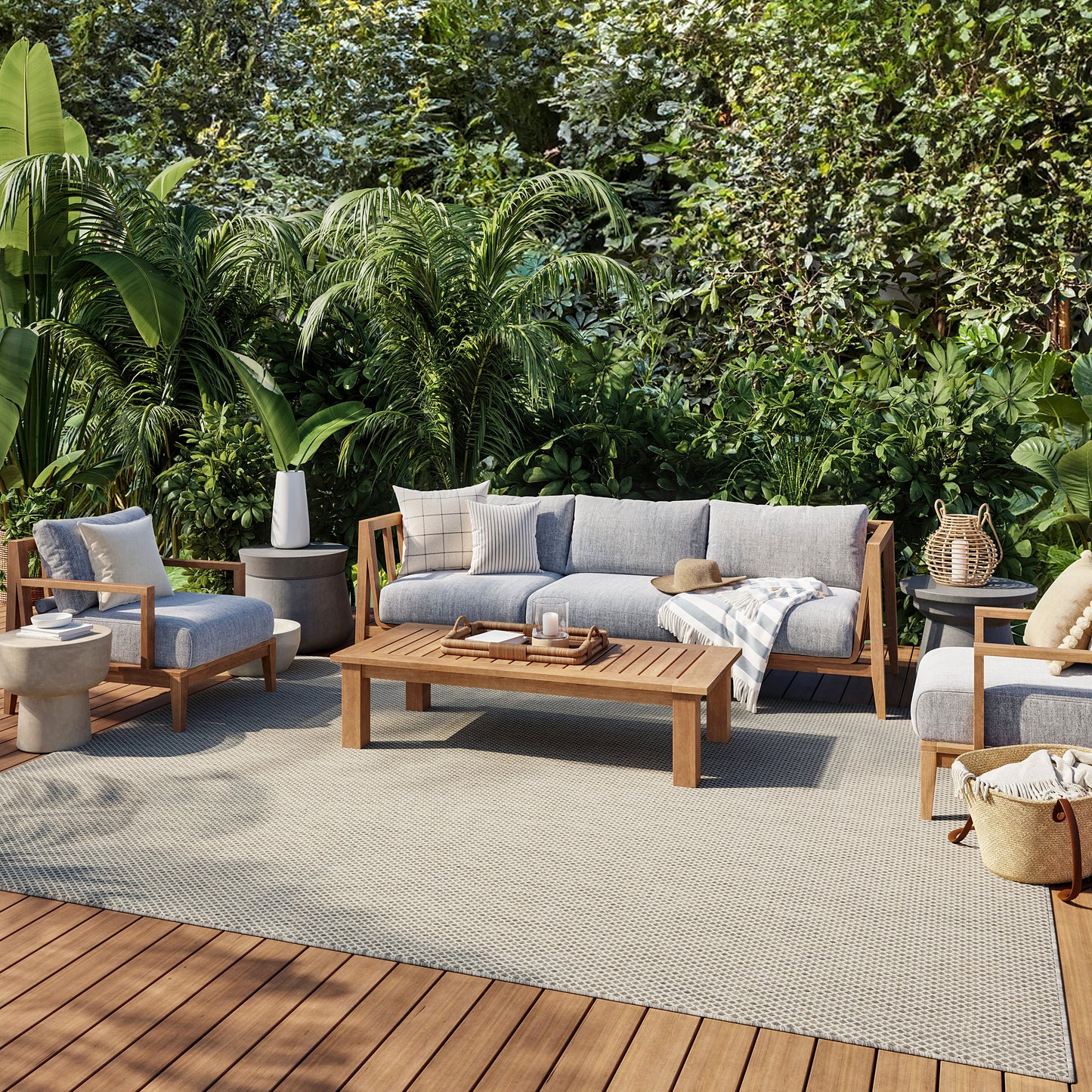 Outdoor wooden-framed patio set with grey cushions, coffee table and chairs on a deck amid lush tropical plants.