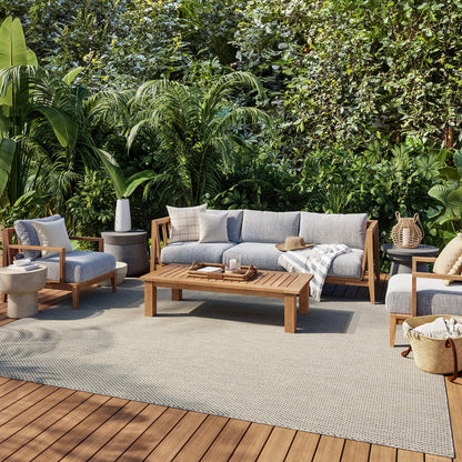 Outdoor wooden-framed patio set with grey cushions, coffee table and chairs on a deck amid lush tropical plants.