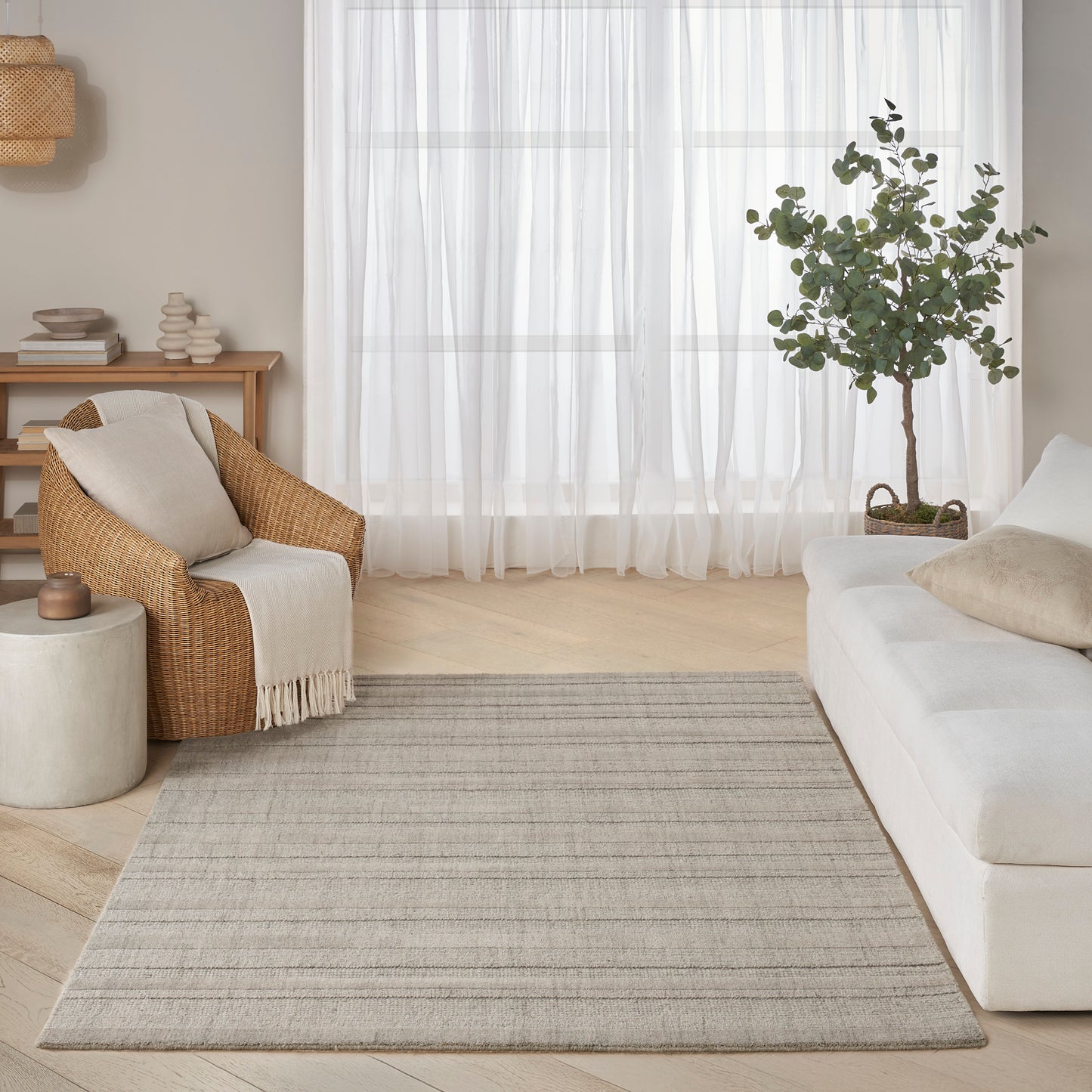 Bright neutral living room with light gray striped woven rug, cream sofa, wicker chair, potted tree and sheer curtains.