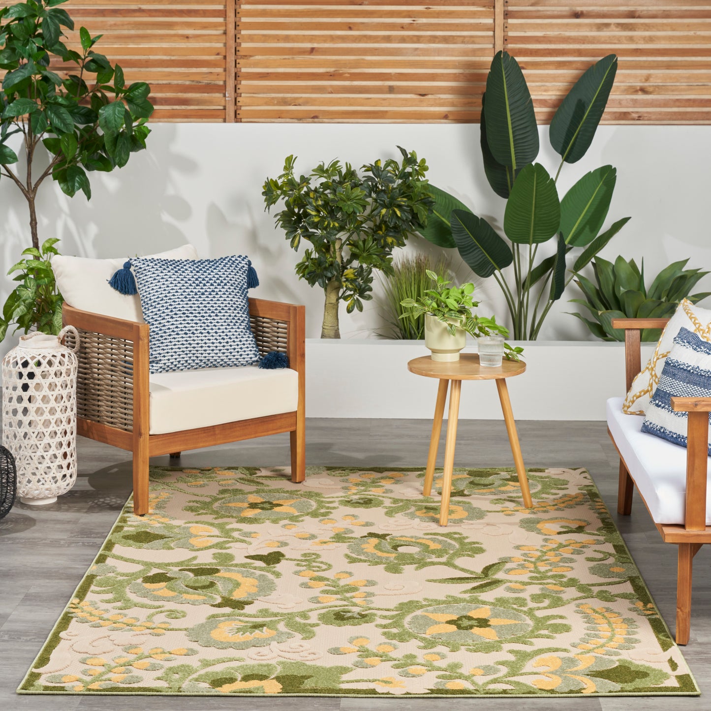 Wood-framed seating with white cushions, blue pillows, round wood table, and green floral rug amid tropical plants.