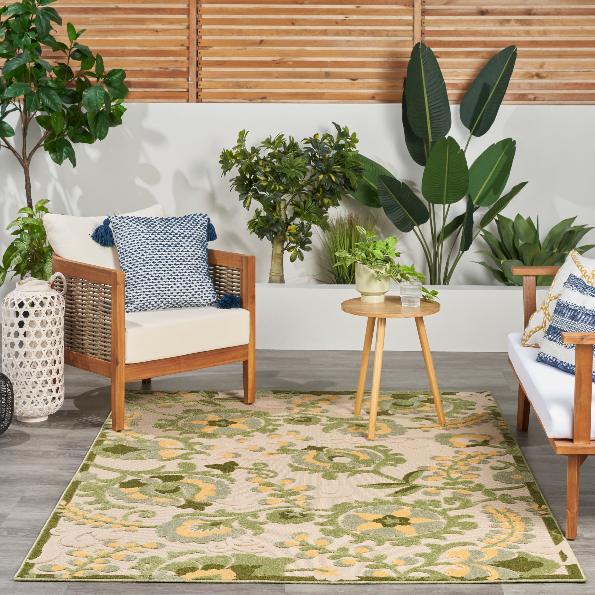 Wood-framed seating with white cushions, blue pillows, round wood table, and green floral rug amid tropical plants.