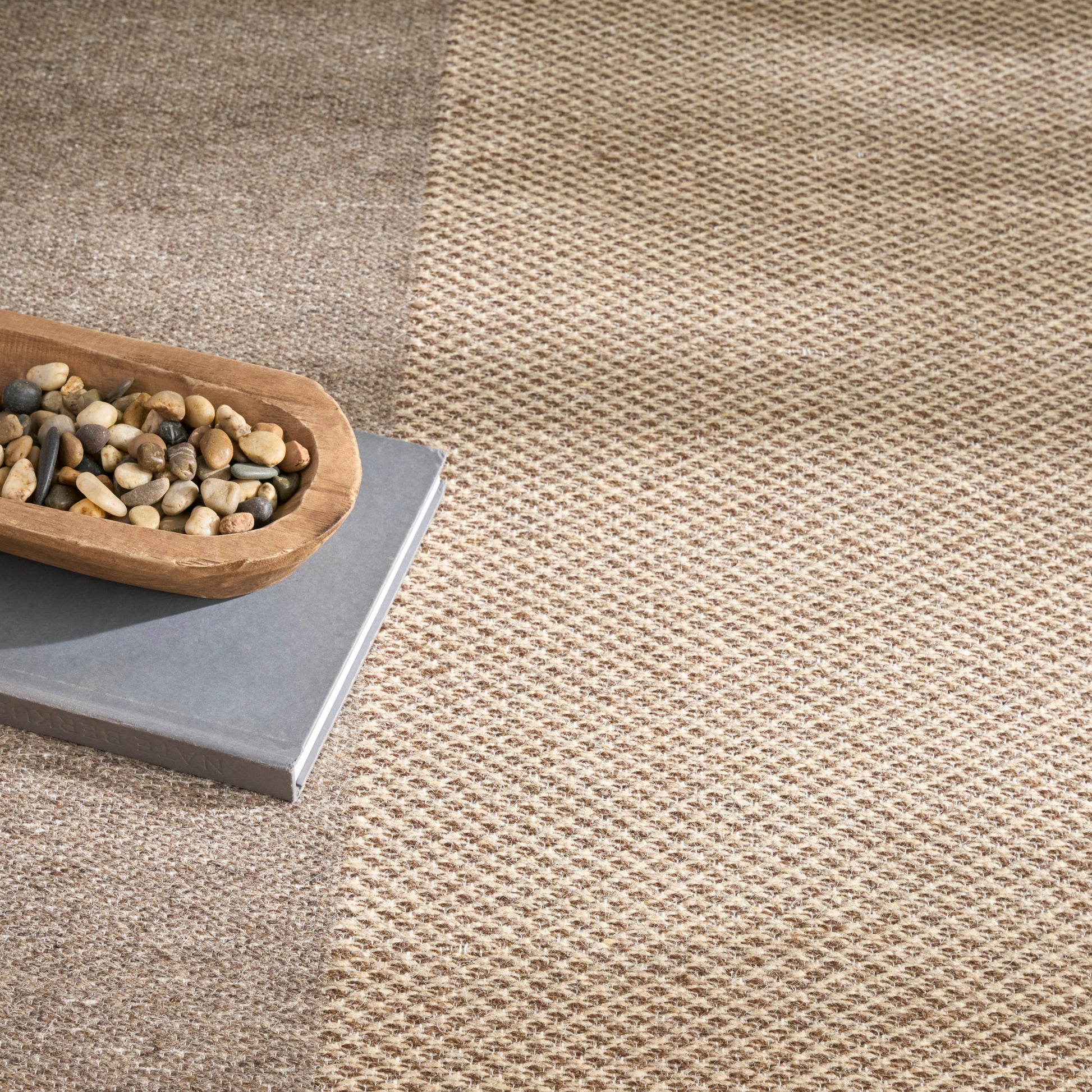 Natural beige woven rug with a wooden boat-shaped bowl of pebbles resting on a gray book.