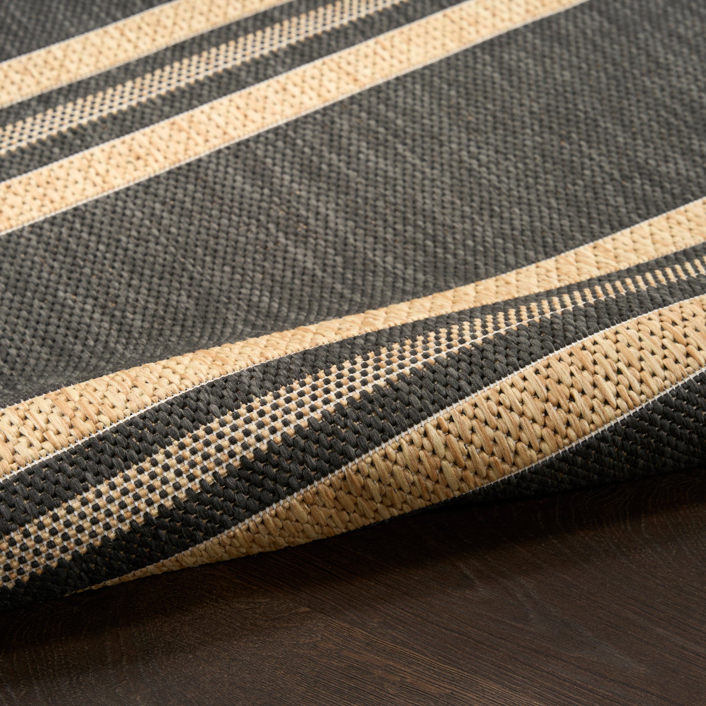 Close-up of a striped woven rug in charcoal gray and natural beige tones, textured fibers, folded on a dark wood surface.